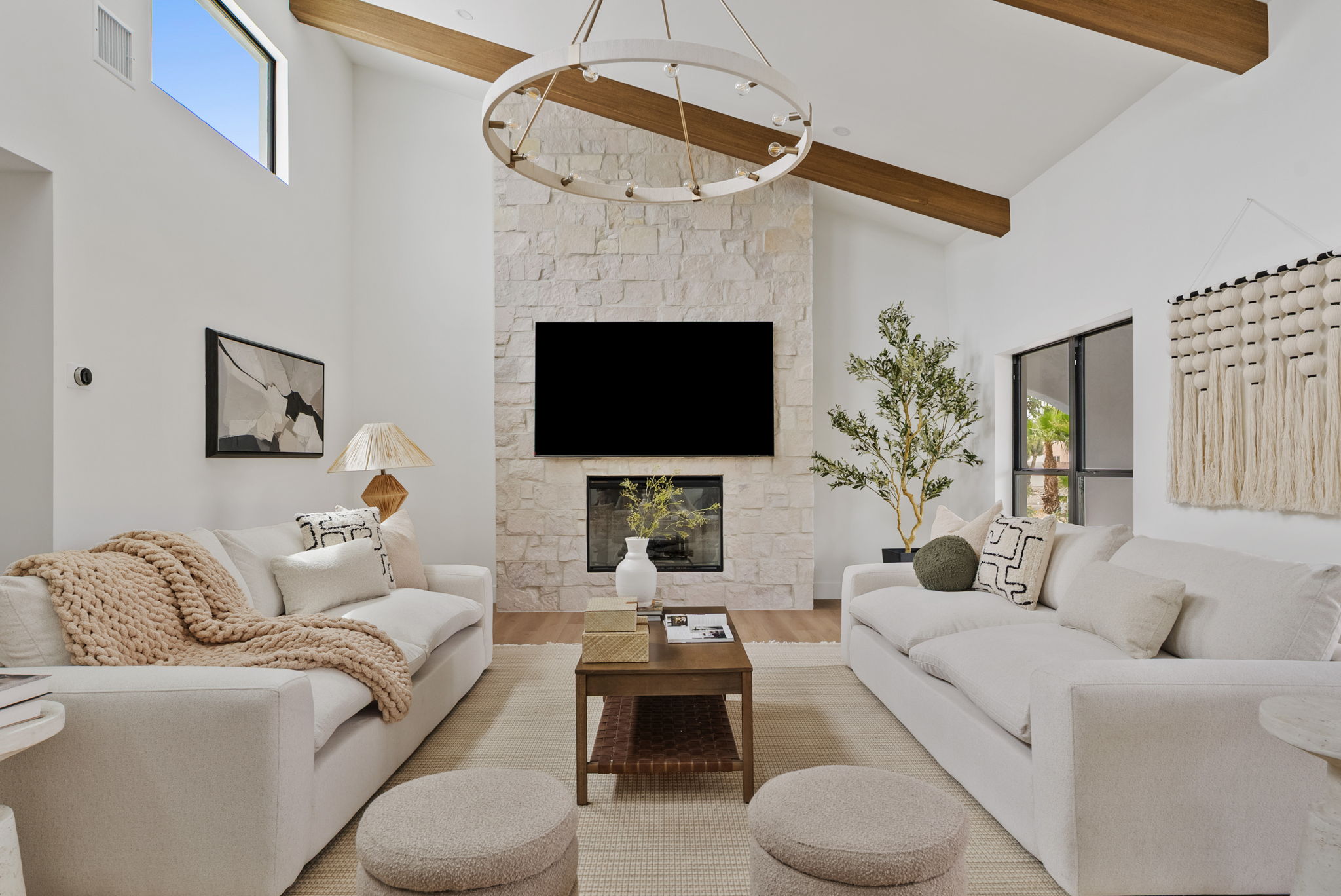 A bright, modern living room features two plush white sofas, a stone fireplace with a mounted television, a coffee table, and a light-colored rug. High vaulted ceilings with wooden beams and a large circular chandelier complete the inviting space.