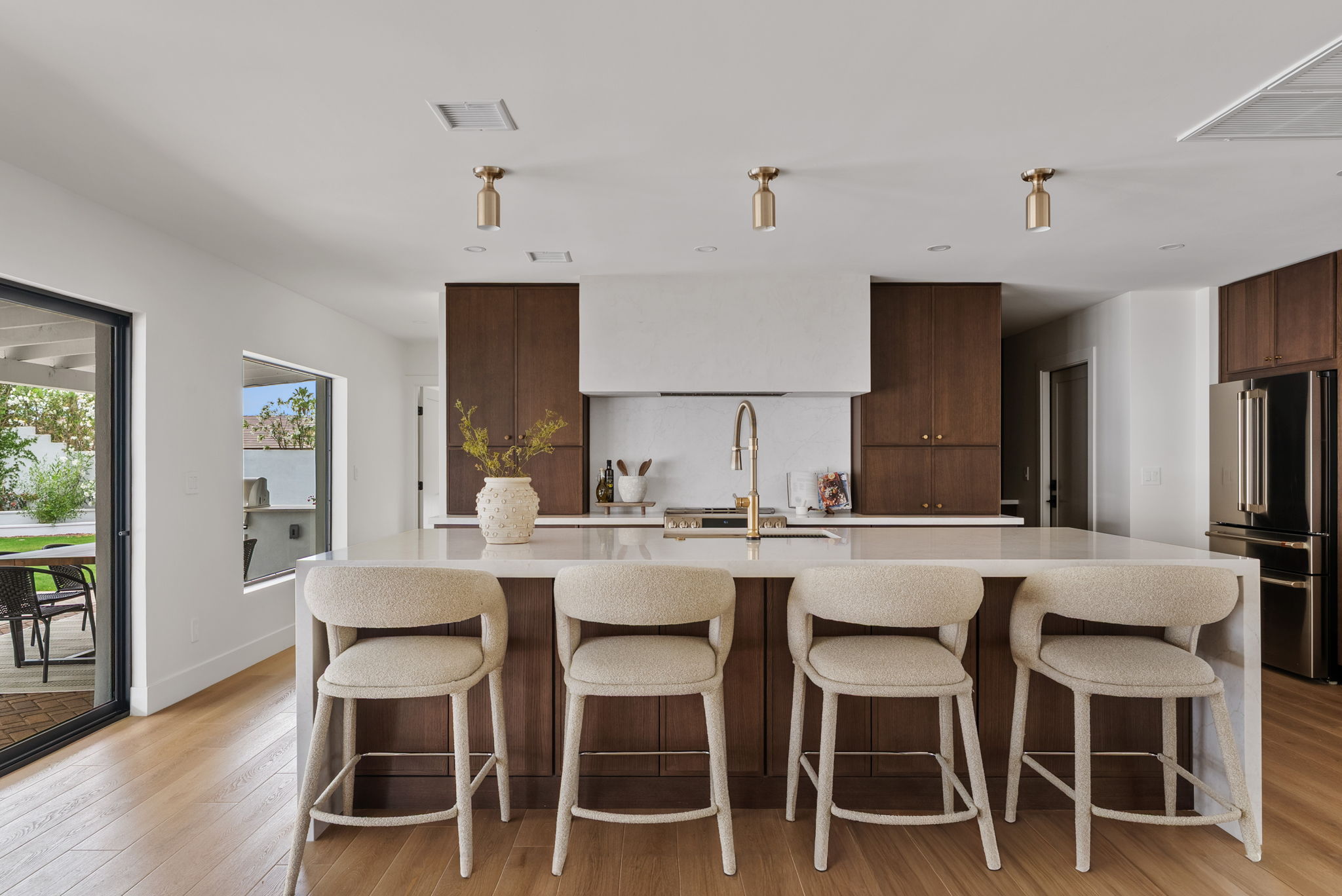 Modern kitchen with a large white island, four light-colored bar stools, dark wood cabinets, and a glass door leading to an outdoor patio.