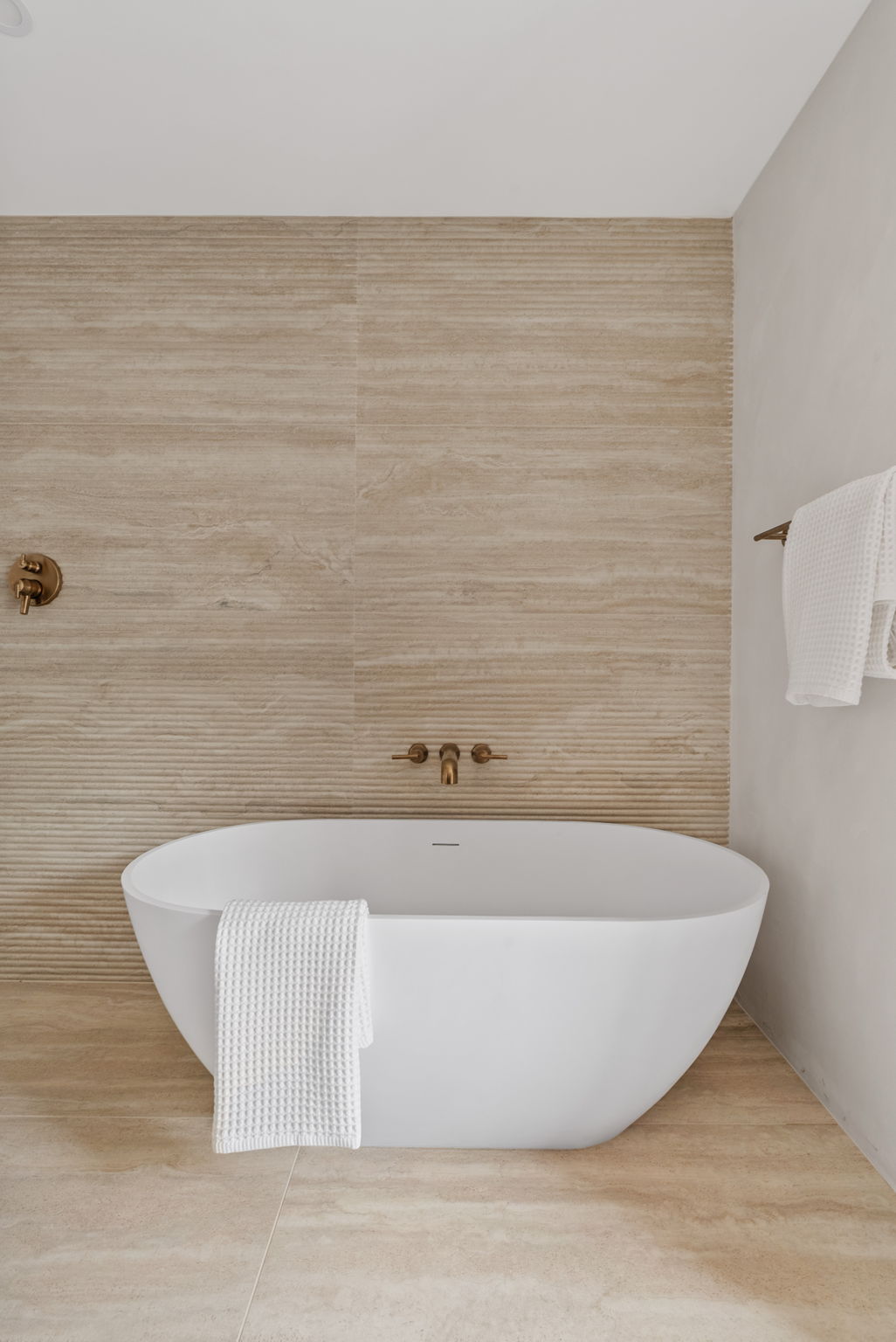 A modern bathroom features a white freestanding bathtub, beige textured wall tiles, bronze fixtures, and white waffle-weave towels.