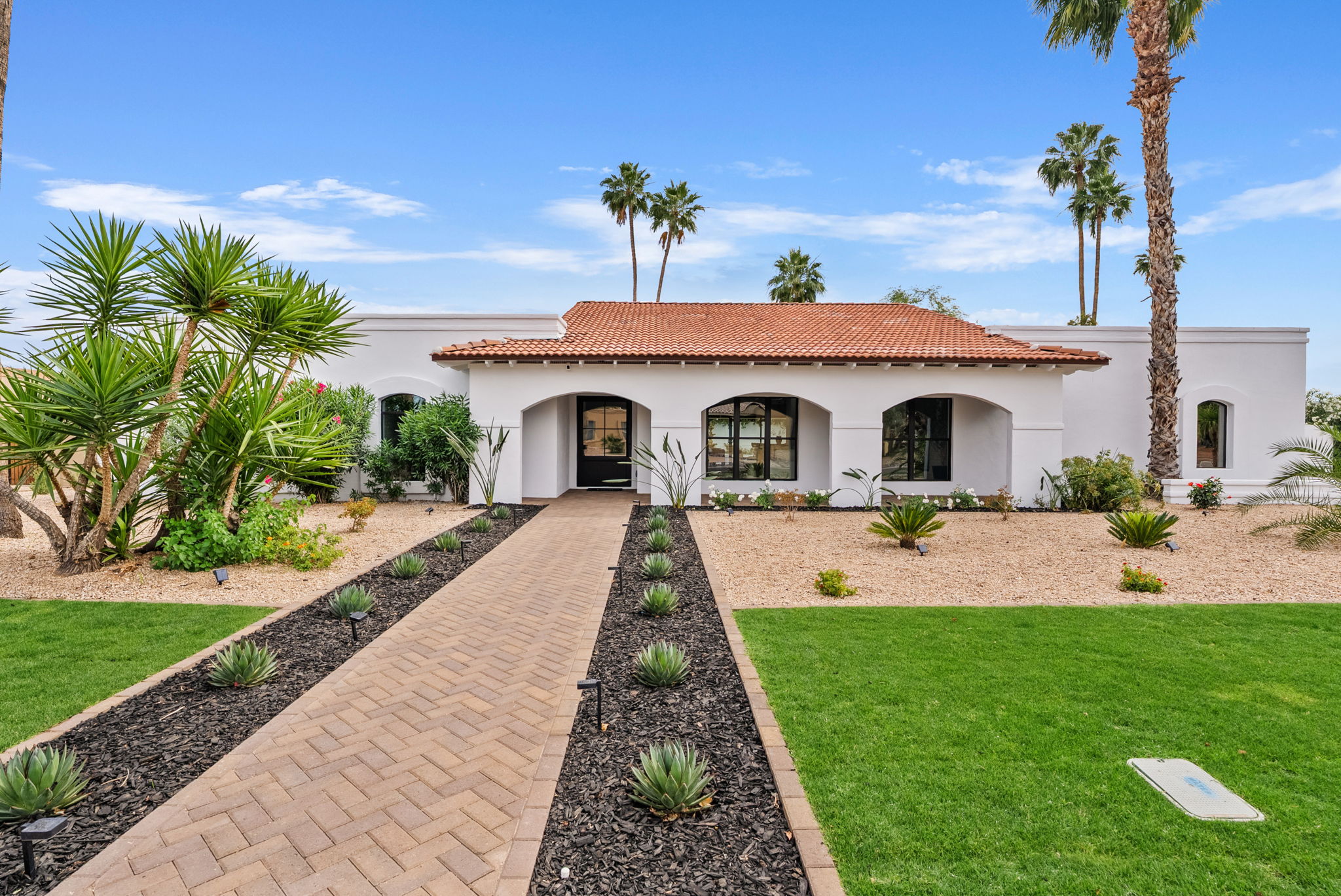 A white single-story Mediterranean-style house with a red tile roof and arched entryways. A brick pathway leads through a landscaped front yard with grass, gravel, agave plants, and tall palm trees under a blue sky.