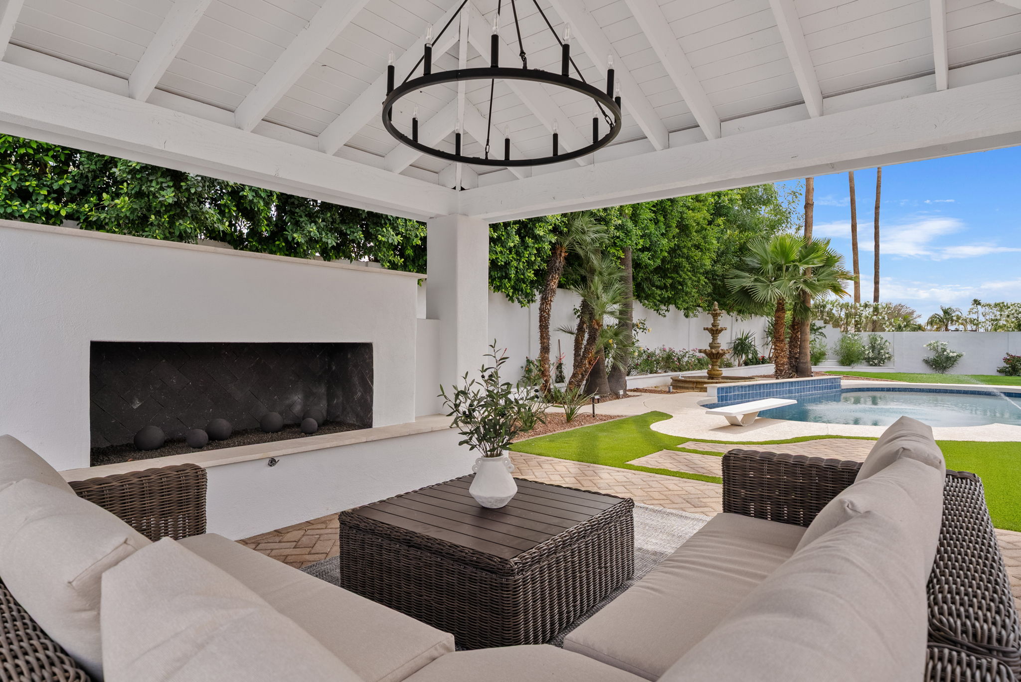 An outdoor patio with wicker seating, a modern fireplace, and a black chandelier under a white pergola, overlooking a pool, fountain, palm trees, and lush green landscaping.