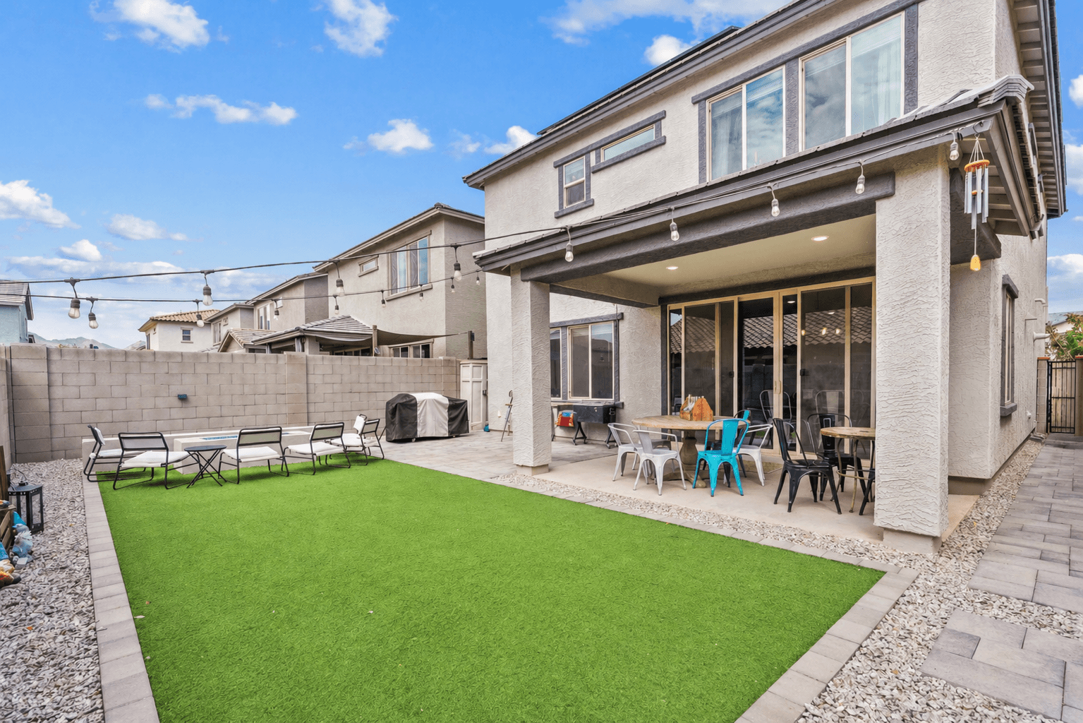 Modern backyard with a two-story house, covered patio, dining and fire pit seating, and artificial turf lawn under string lights.
