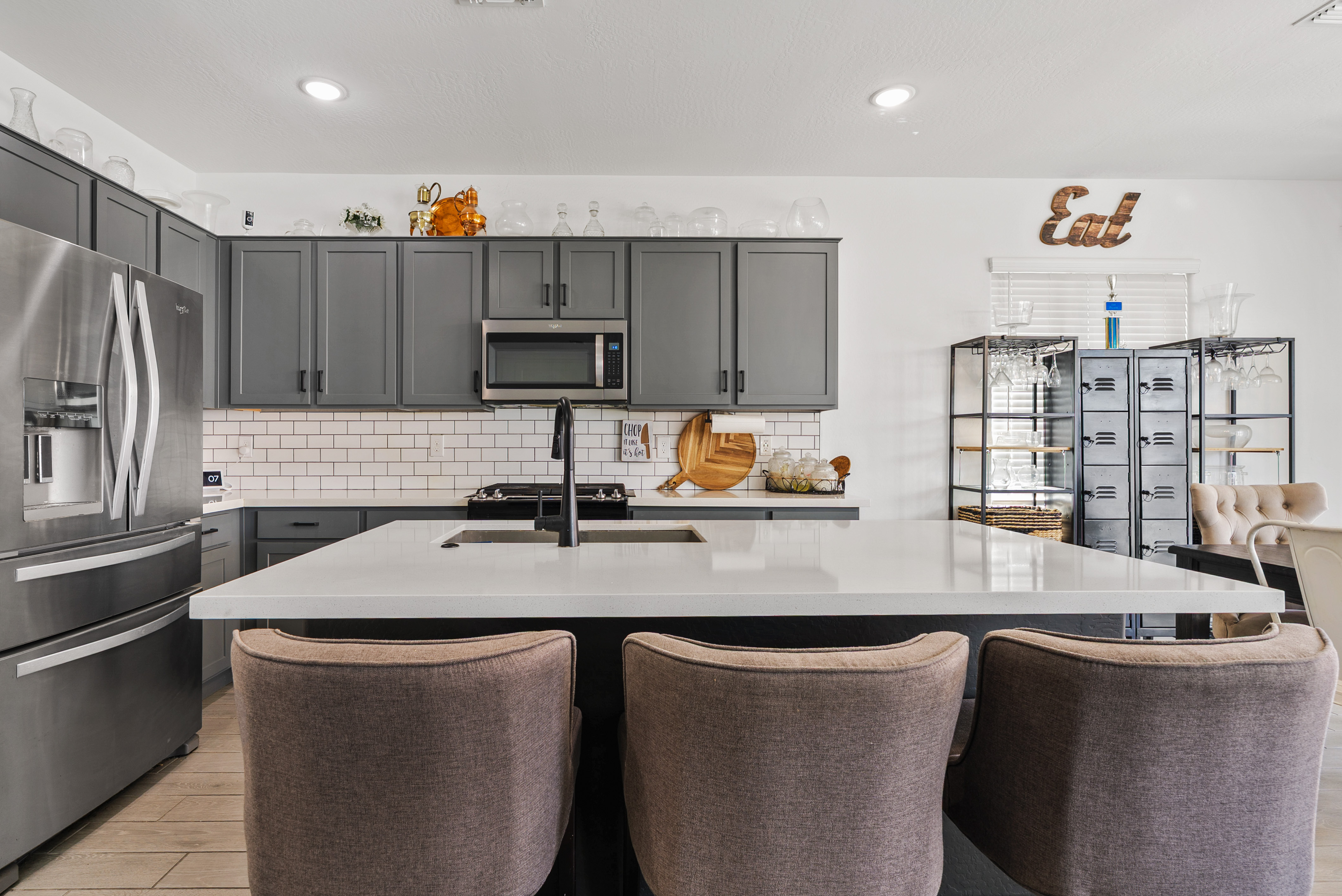 Modern kitchen featuring dark gray cabinets, white subway tile backsplash, stainless steel appliances, a large island with three bar stools, and a wooden 'Eat' sign.