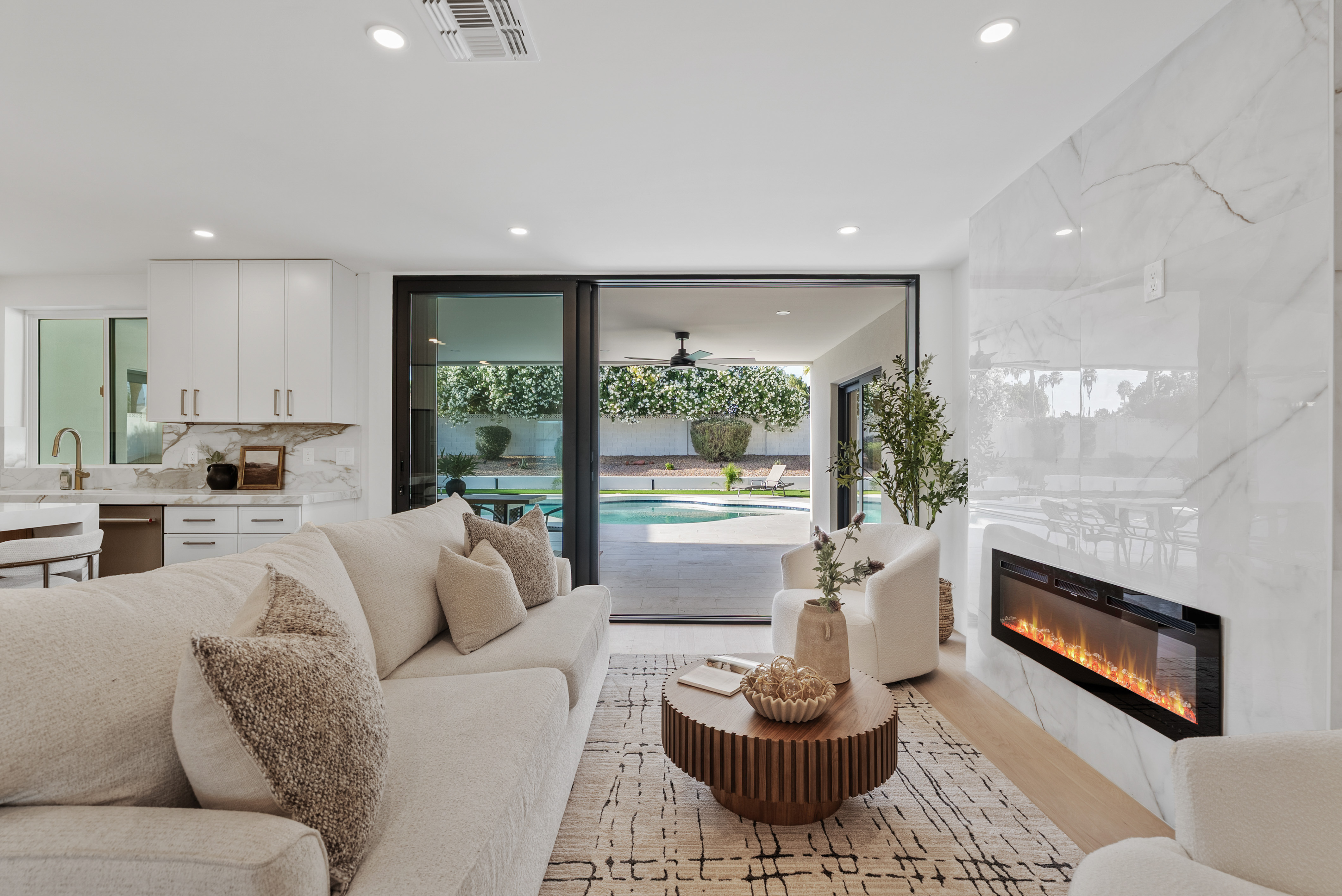 Bright, modern open-concept living room and kitchen with a light sofa, patterned rug, and marble fireplace, opening onto a backyard with a swimming pool.
