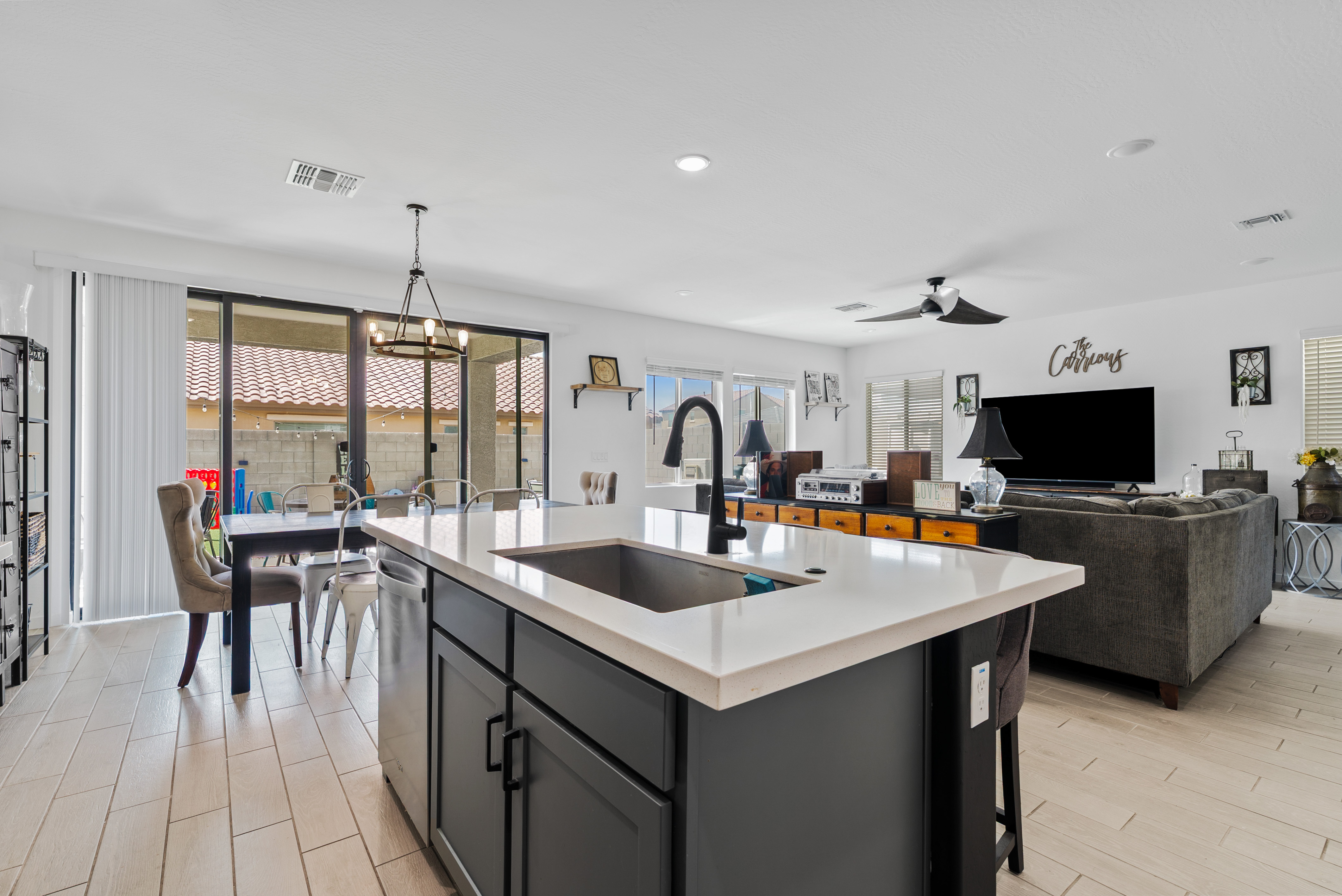 A spacious open-concept living space with a dark kitchen island, light countertops, a dining area, and a living room featuring a large TV, dark sectional sofa, and light tiled floors.