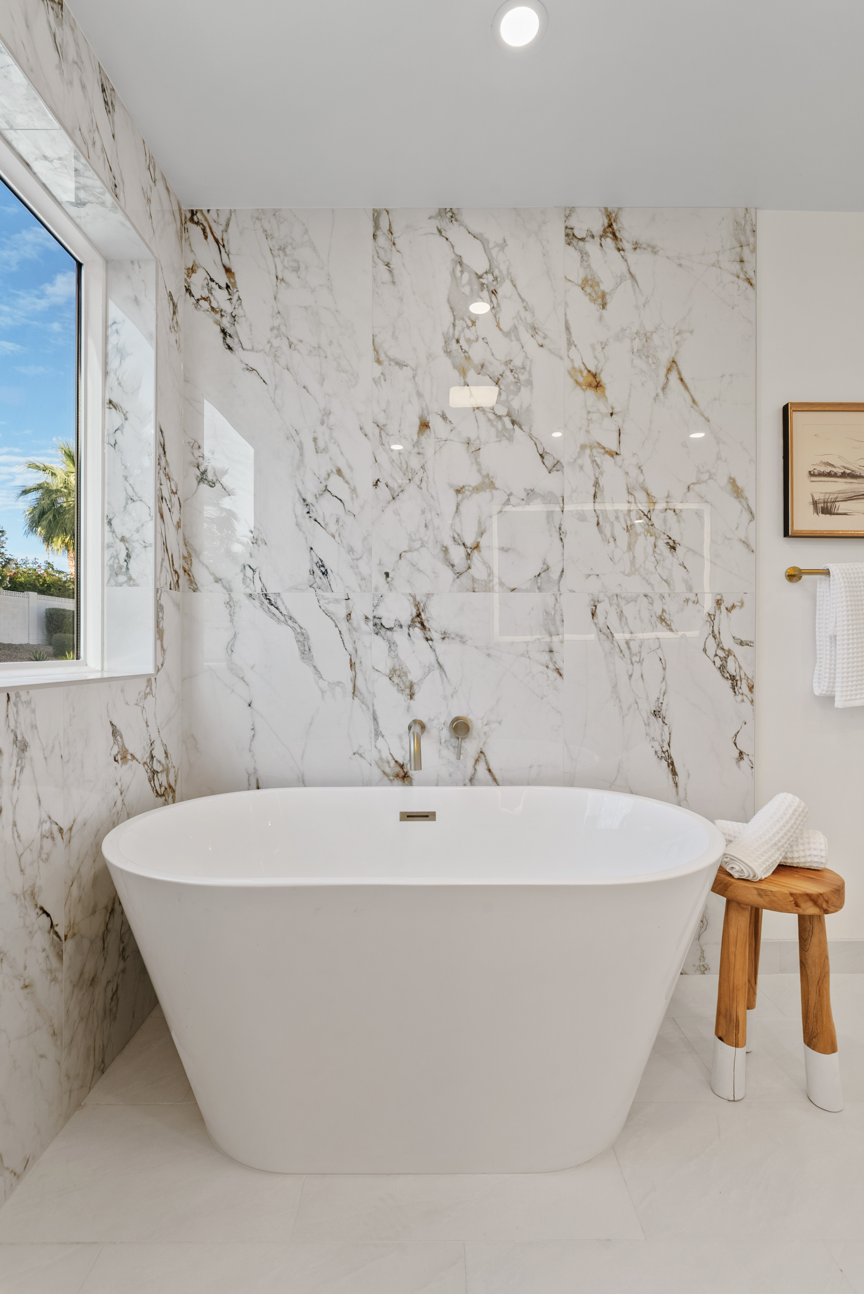 Modern bathroom featuring a freestanding white bathtub against a wall of white marble with gold veins, a large window, and a wooden stool with rolled towels.