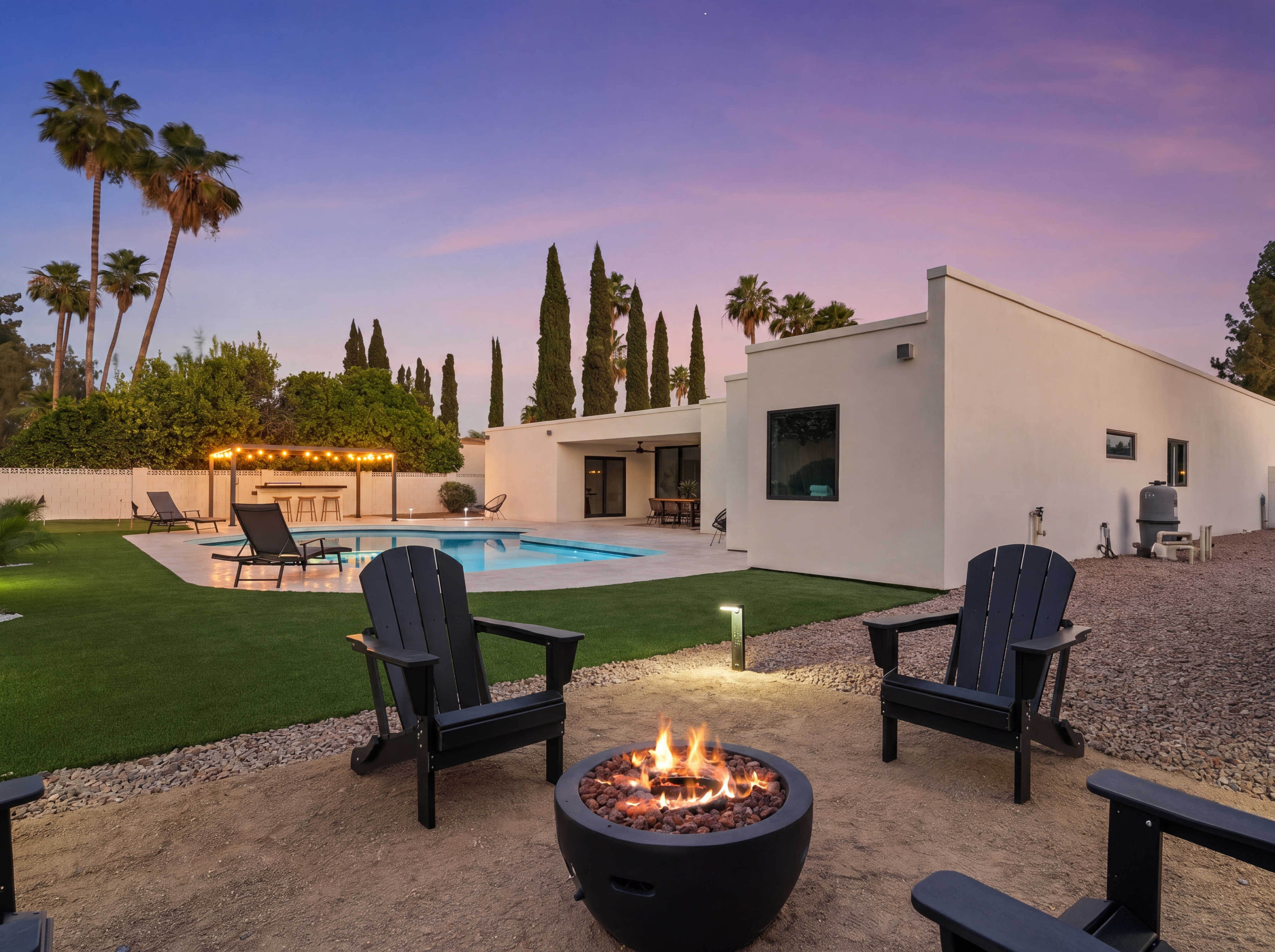 Modern backyard at dusk with a swimming pool, a lit fire pit and Adirondack chairs, a bar area, and a white house under a vibrant sunset sky.