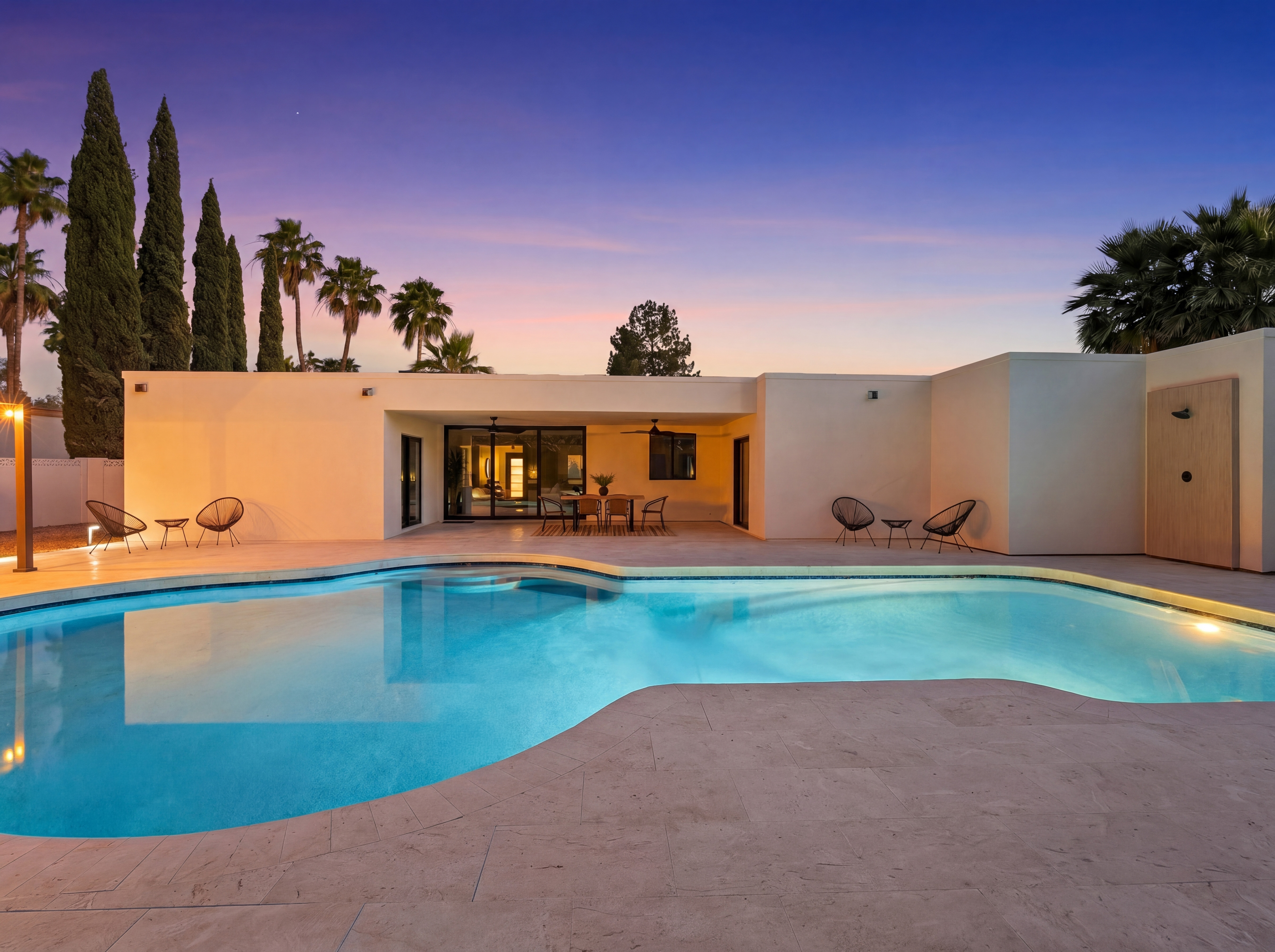 Modern house with a large illuminated swimming pool and patio at twilight, featuring a vibrant gradient sky and palm trees.