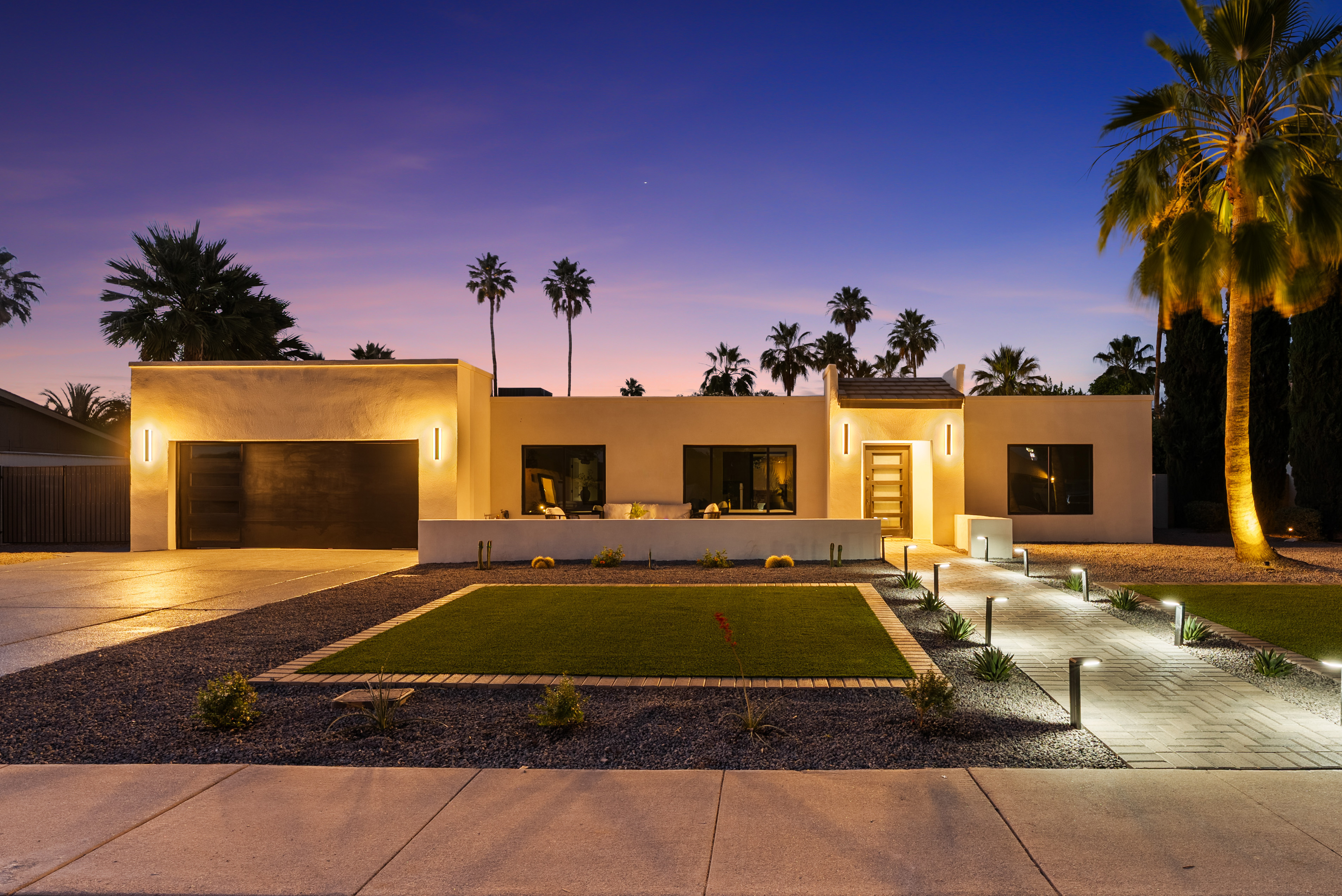 Modern house exterior at twilight with warm lights illuminating the facade, driveway, and a pathway lined with ground lights. Palm trees are visible against the purple and blue sky.
