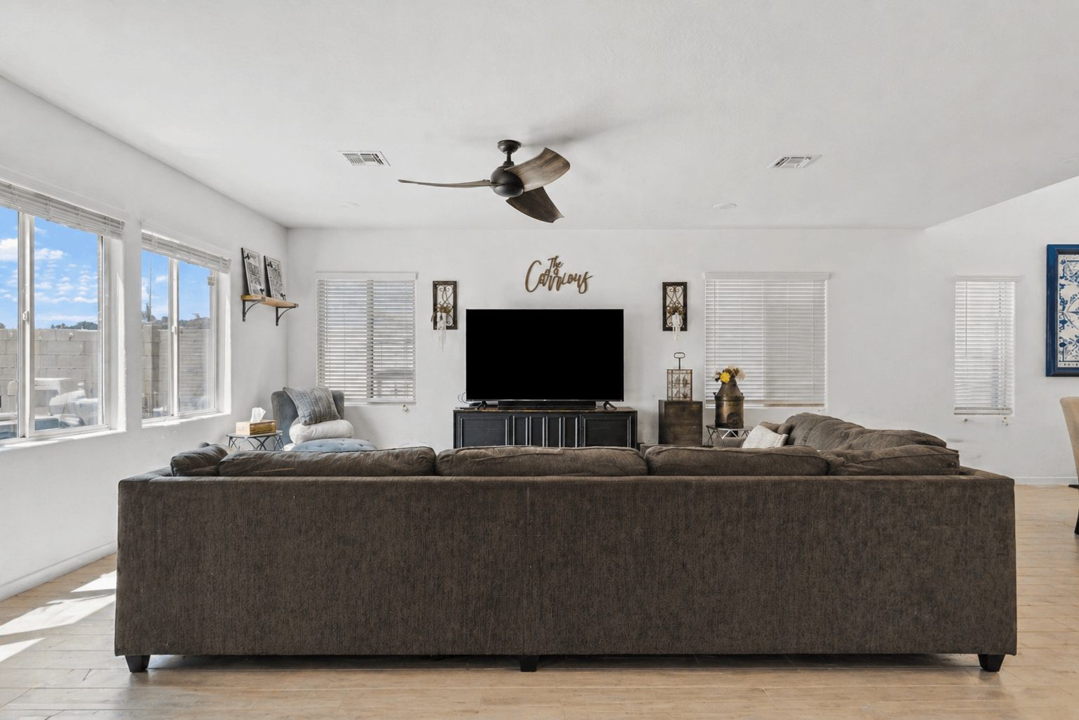 A living room with a large brown sectional sofa, a wall-mounted TV over a credenza, and a wooden "The Carriollas" sign above it.