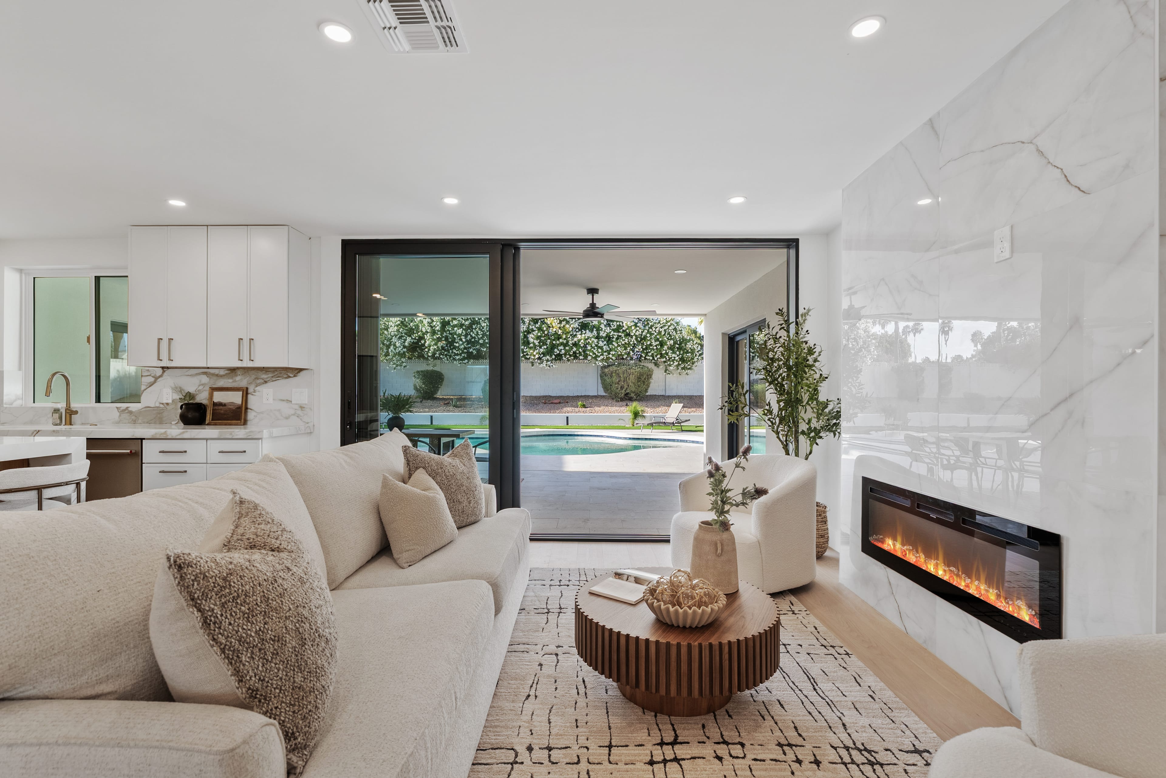 A bright, open-concept living room features a cream sectional, a round wood coffee table, and a marble fireplace wall, with sliding doors leading to a backyard pool.