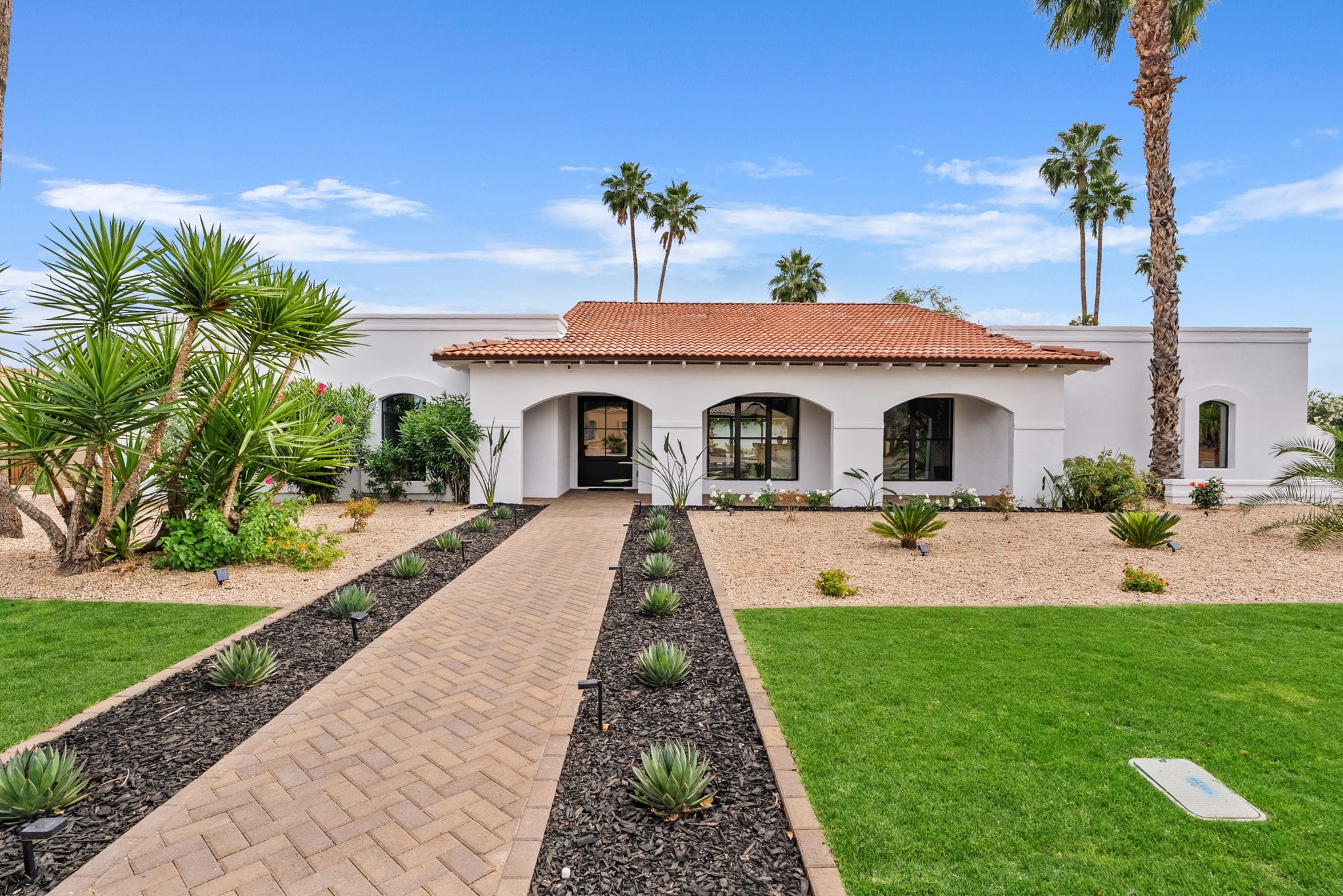 A Mediterranean-style white stucco house with a red tile roof, arched entry, and windows, surrounded by a neatly landscaped yard with a brick path, green lawn, and various palm trees.