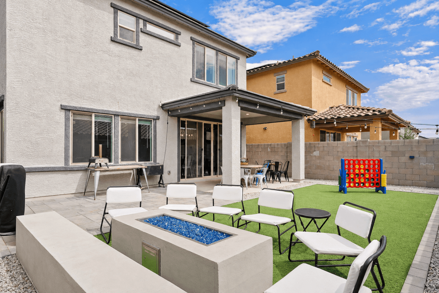 A residential backyard featuring a fire pit with blue glass, white chairs on artificial grass, a covered patio, and a large Connect 4 game.