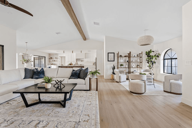 A modern, open-concept living room and kitchen with a white sectional sofa, dark coffee table, and a separate seating area with accent chairs and a tall bookshelf, all under a vaulted ceiling.