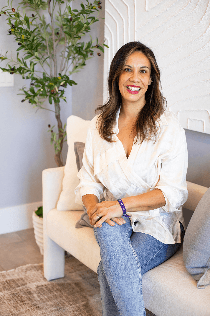 A friendly woman with long brown hair smiles at the camera, sitting casually on a cream couch in a modern room with a plant and textured wall art.
