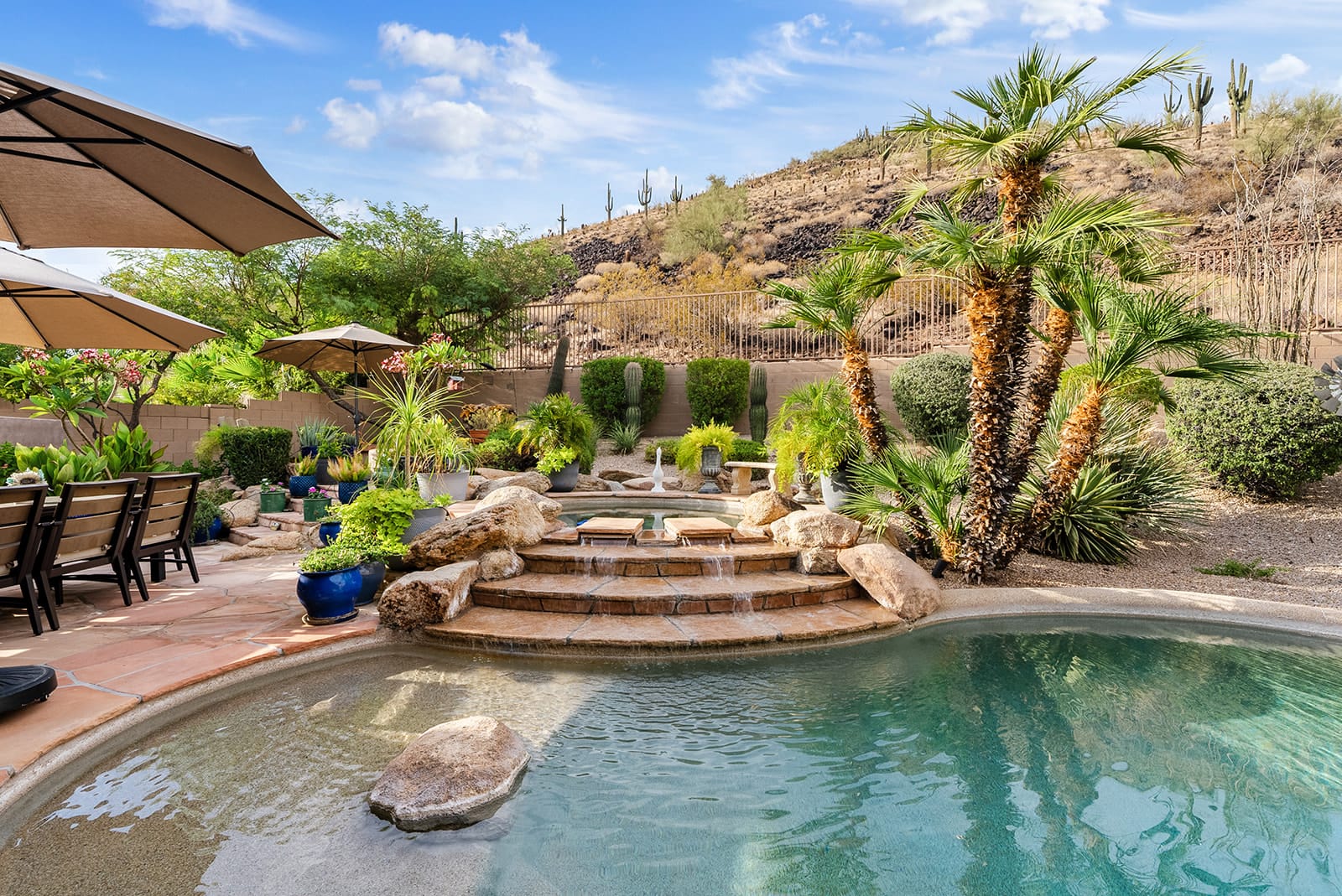 Luxurious backyard with a swimming pool, tiered hot tub, outdoor dining area, and lush desert landscaping featuring palm trees and cacti, against a desert hillside.
