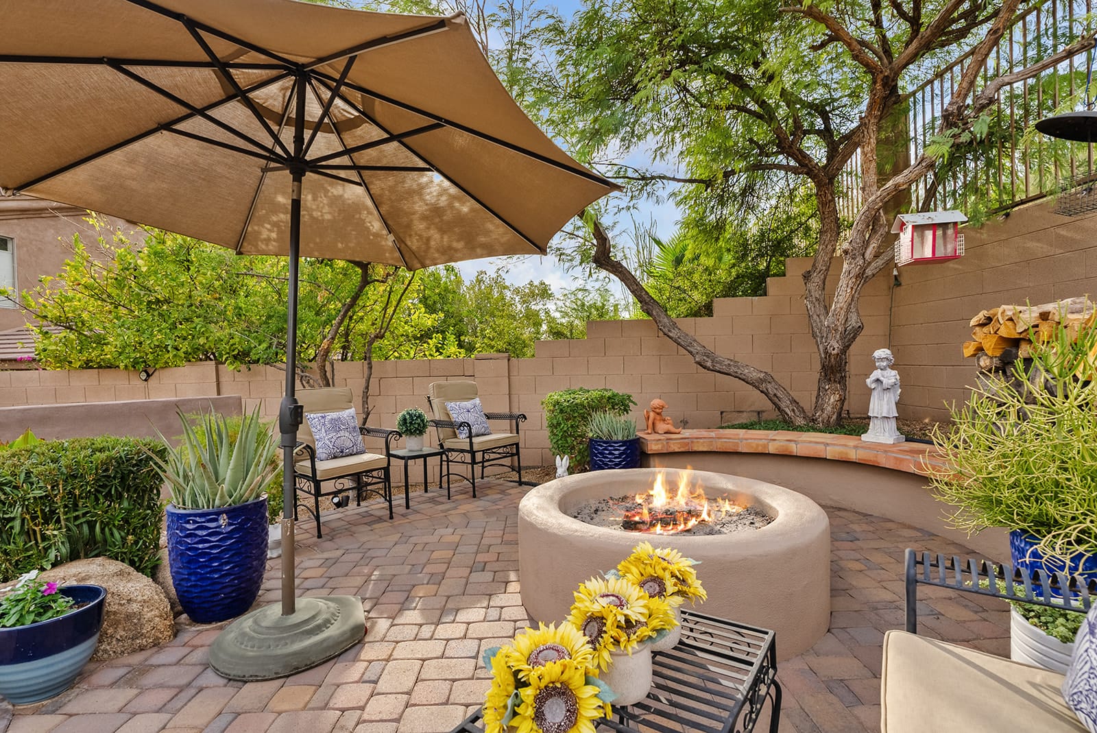 An inviting outdoor patio features a central lit fire pit, comfortable chairs, potted sunflowers, and an open umbrella, set against a retaining wall with green trees.