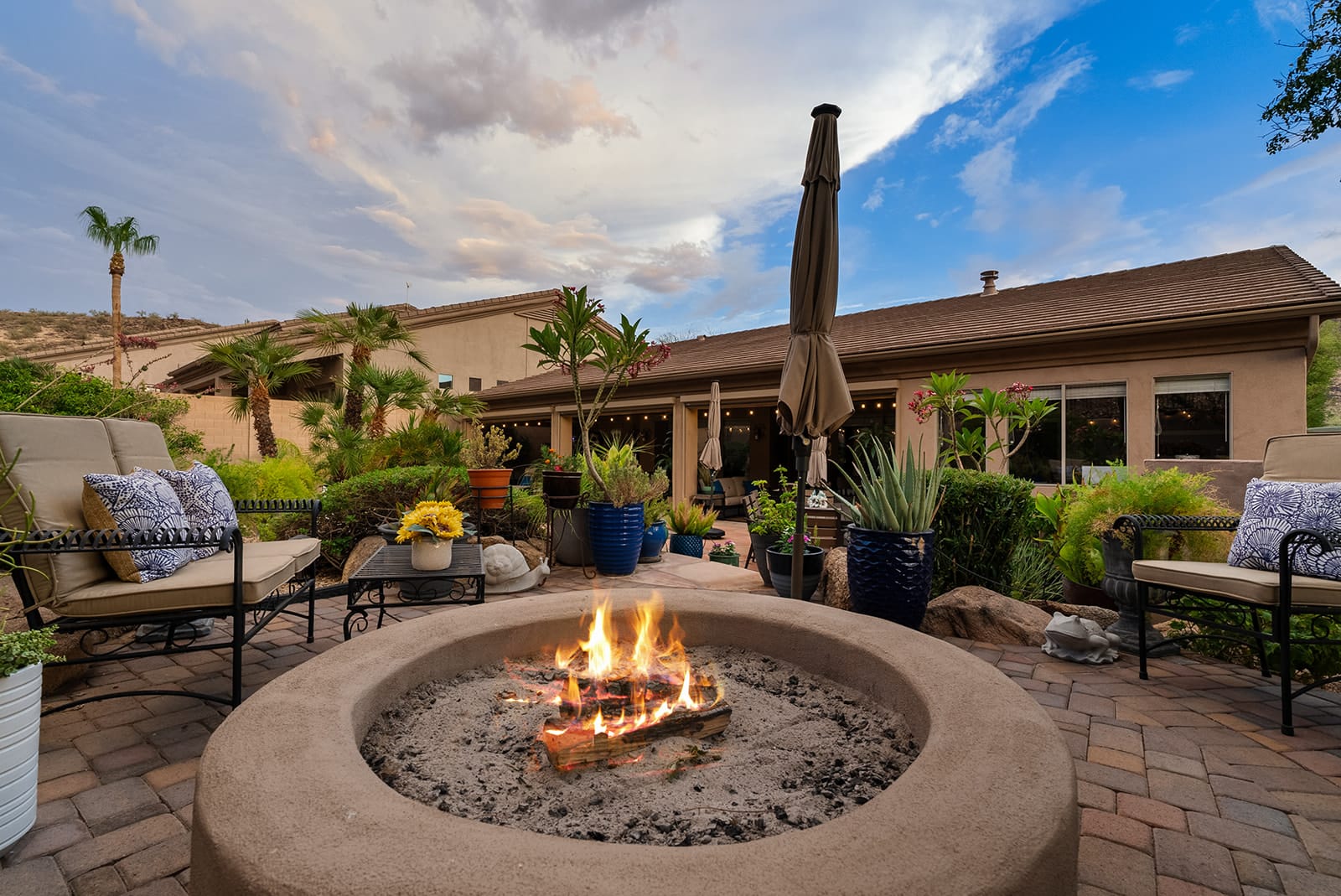 A cozy outdoor patio at dusk with a lit fire pit, comfortable seating, potted plants, and a house in the background under a cloudy sky.