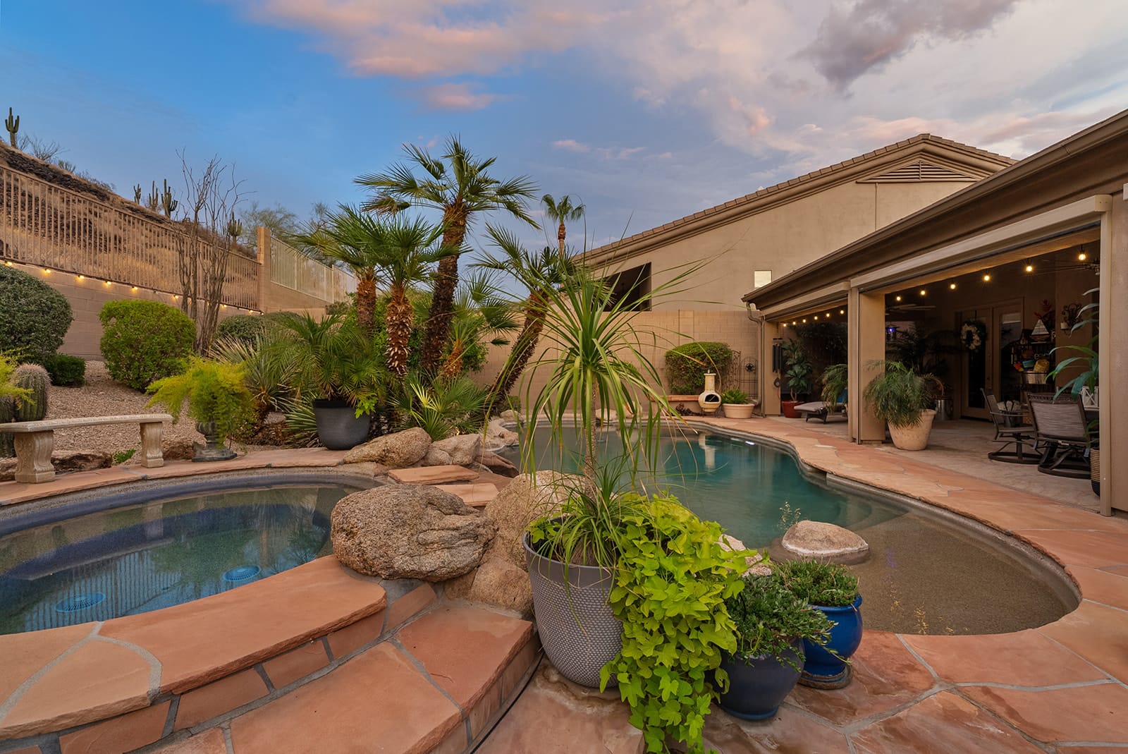 A beautifully lit desert backyard at dusk, featuring a swimming pool, hot tub, lush landscaping with palm trees and cacti, and a patio with string lights.