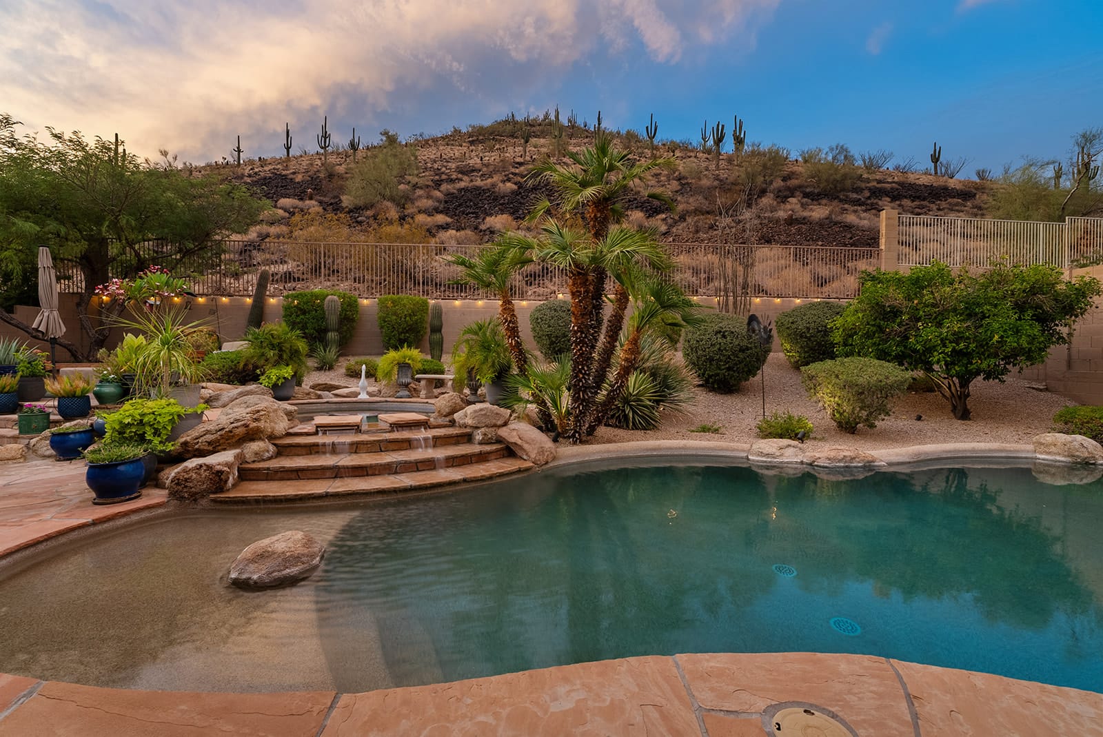 Desert backyard with a swimming pool, tiered waterfall, and lush potted plants, set against a cactus-covered hill under a twilight sky.