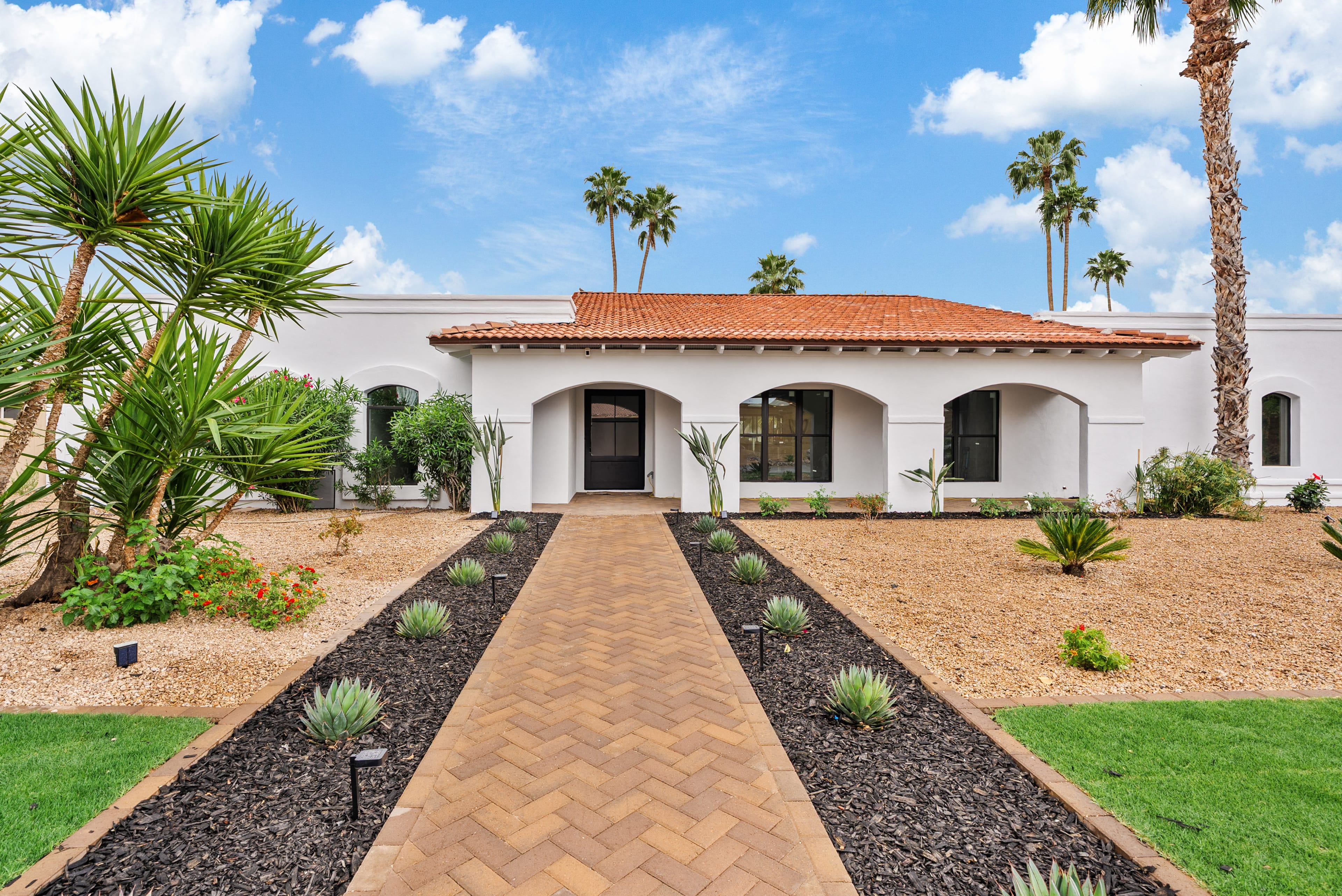 A white, single-story house with a red tile roof and arched entryways, featuring desert landscaping with a brick walkway and palm trees under a blue sky.