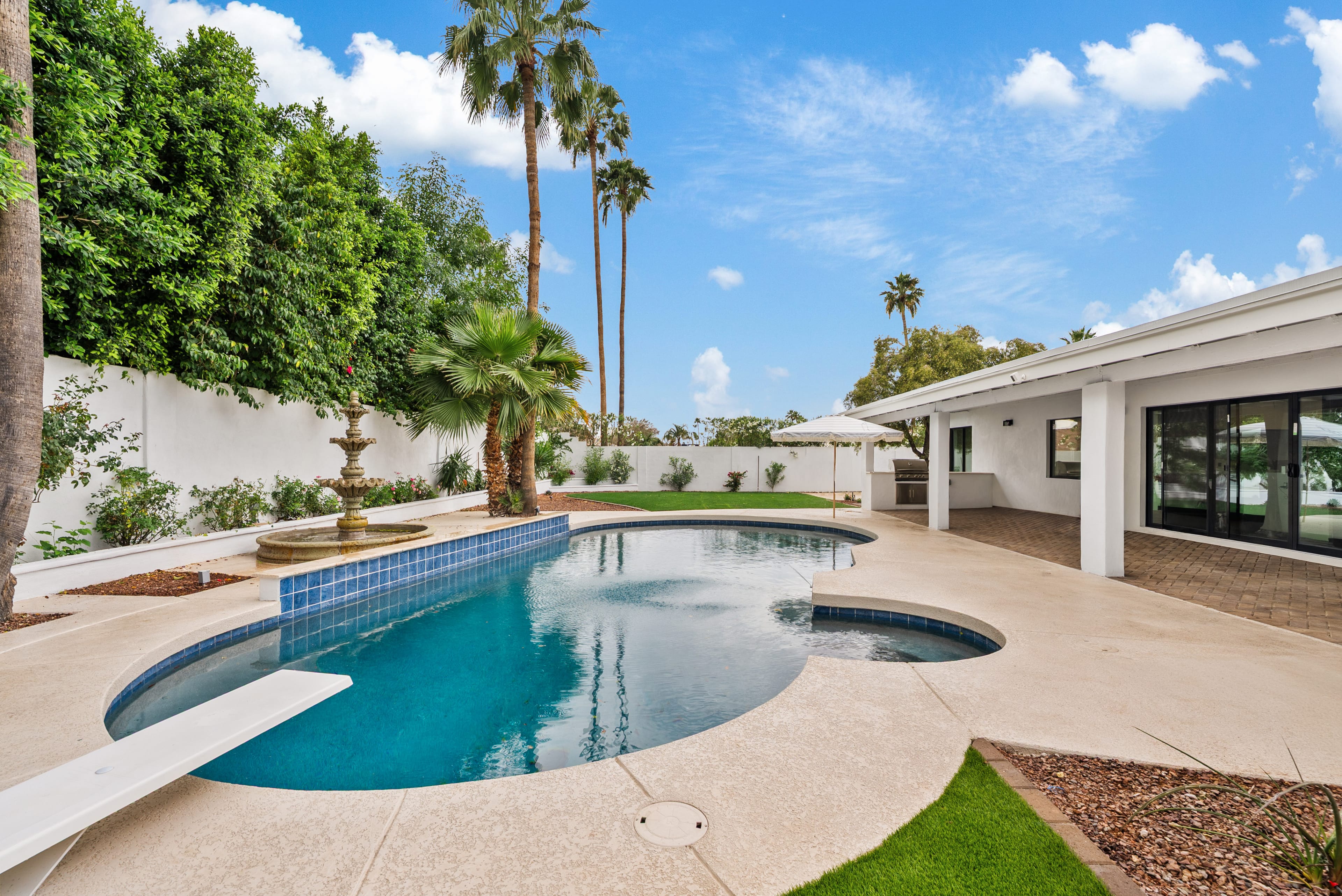 A well-maintained backyard features a blue swimming pool with a diving board, a stone fountain, and an outdoor patio with a BBQ grill. Palm trees and lush green plants line a white wall under a partly cloudy sky.