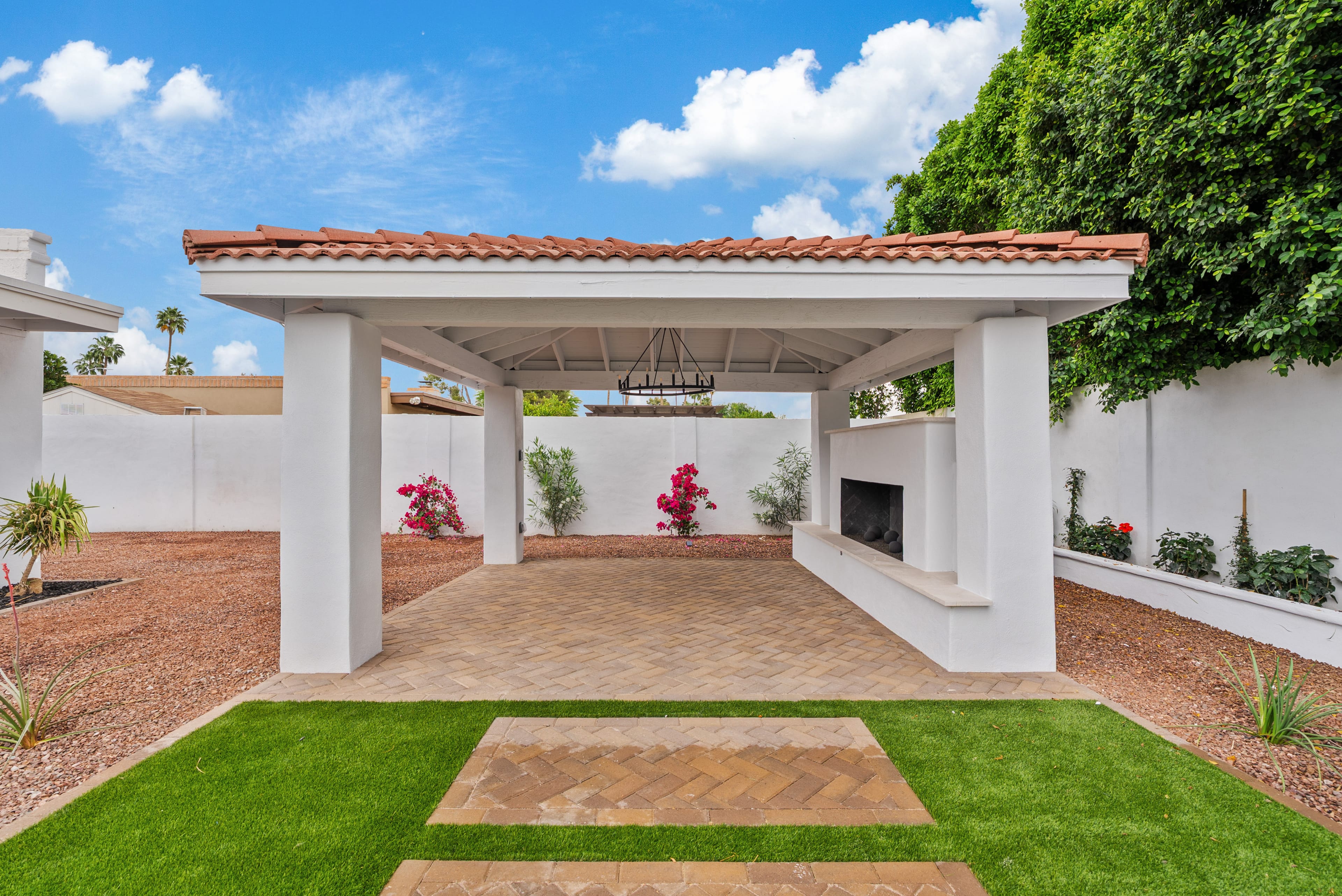 Backyard with a white pergola covering a brick patio, an outdoor fireplace, artificial turf, and gravel landscaping under a blue sky.