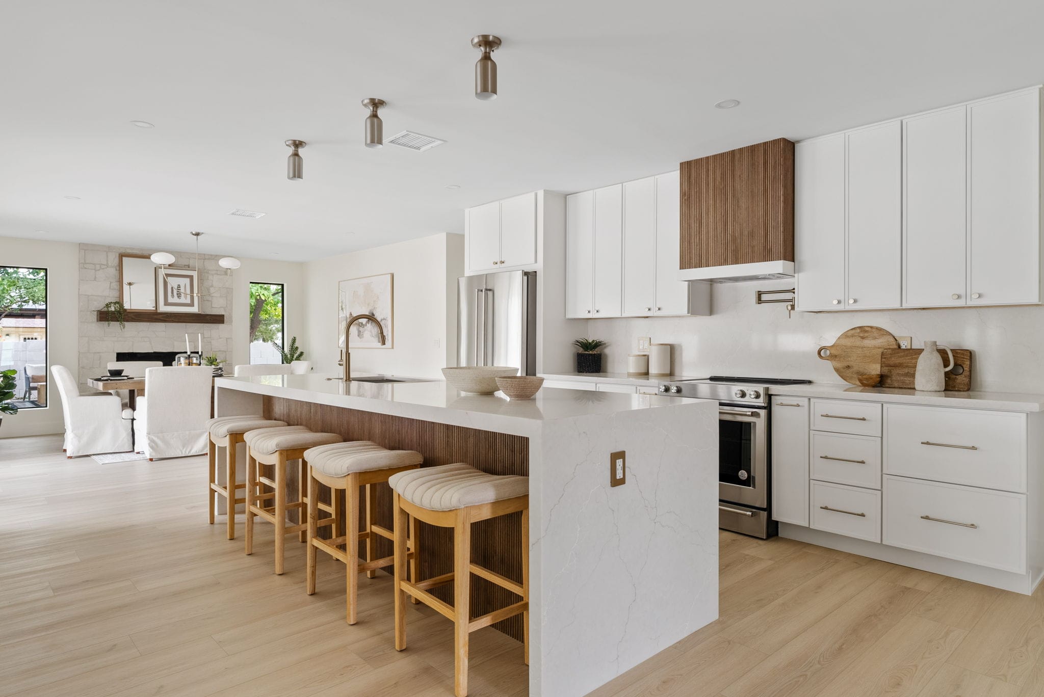 A modern open-concept kitchen and dining area with white cabinets, a large island with bar stools, light wood flooring, and a stone fireplace.