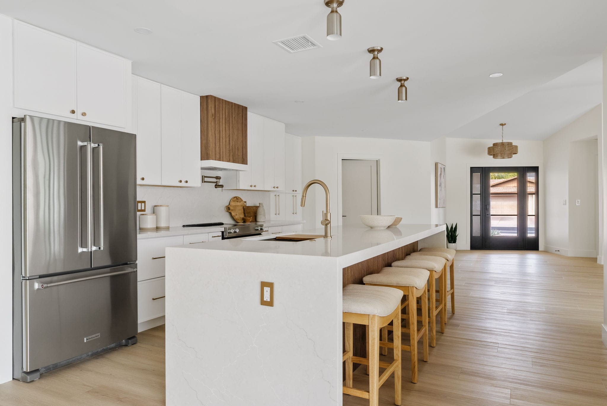 A modern open-concept kitchen with white cabinets, stainless steel appliances, a large island with four stools, brass fixtures, and light wood flooring.