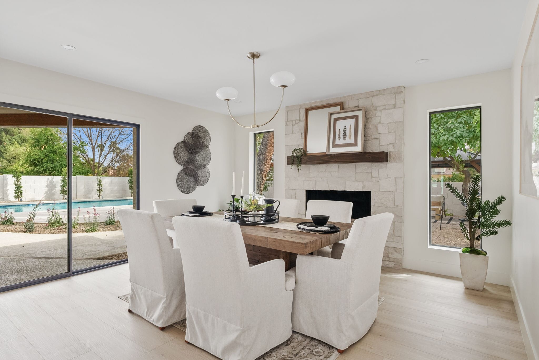 Modern dining room with a large wooden table, white slipcovered chairs, a stone fireplace, and sliding glass doors revealing a swimming pool.