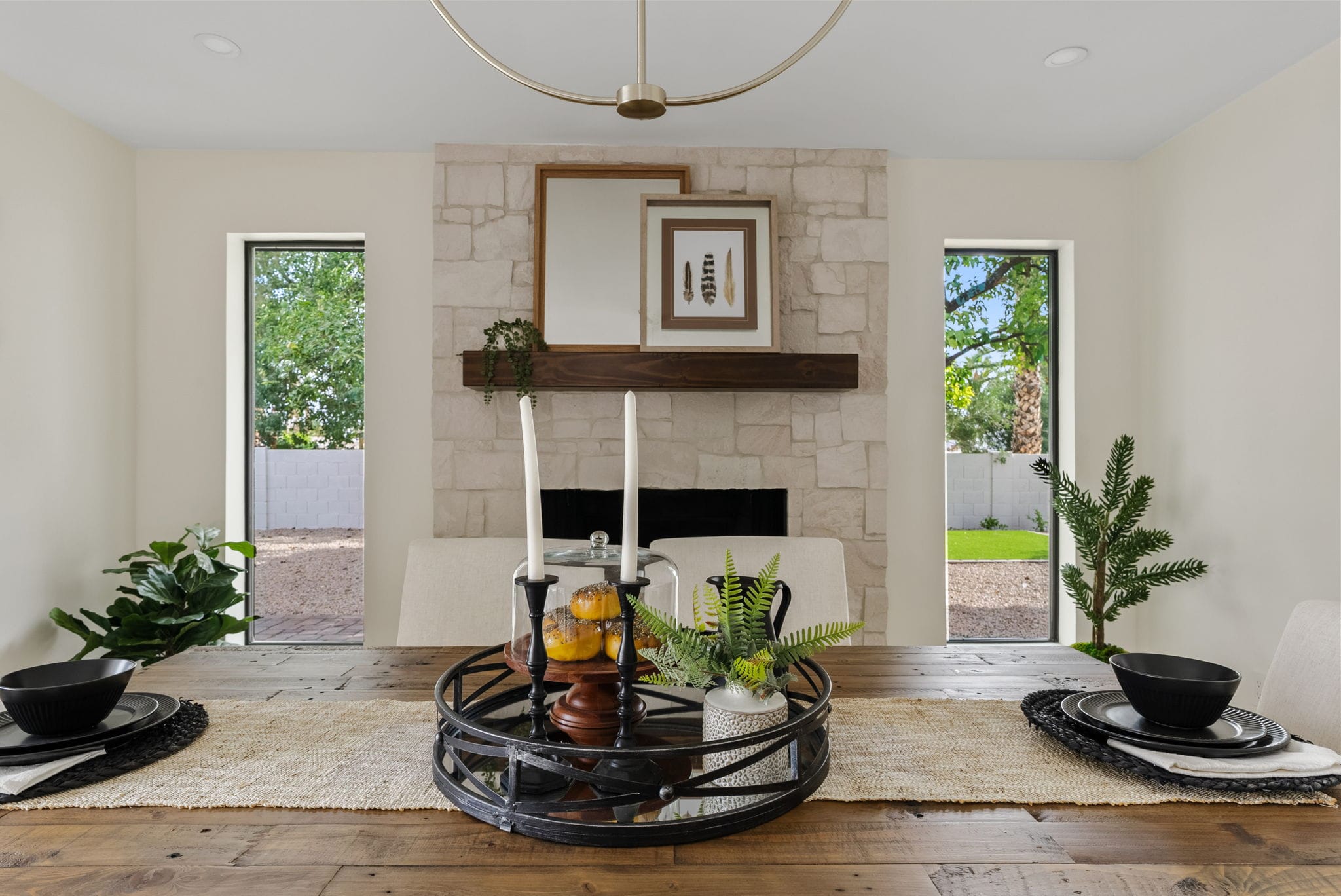 A dining room with a rustic wooden table, set with black dishes and a centerpiece tray, facing a stone fireplace flanked by windows revealing a green outdoor view.