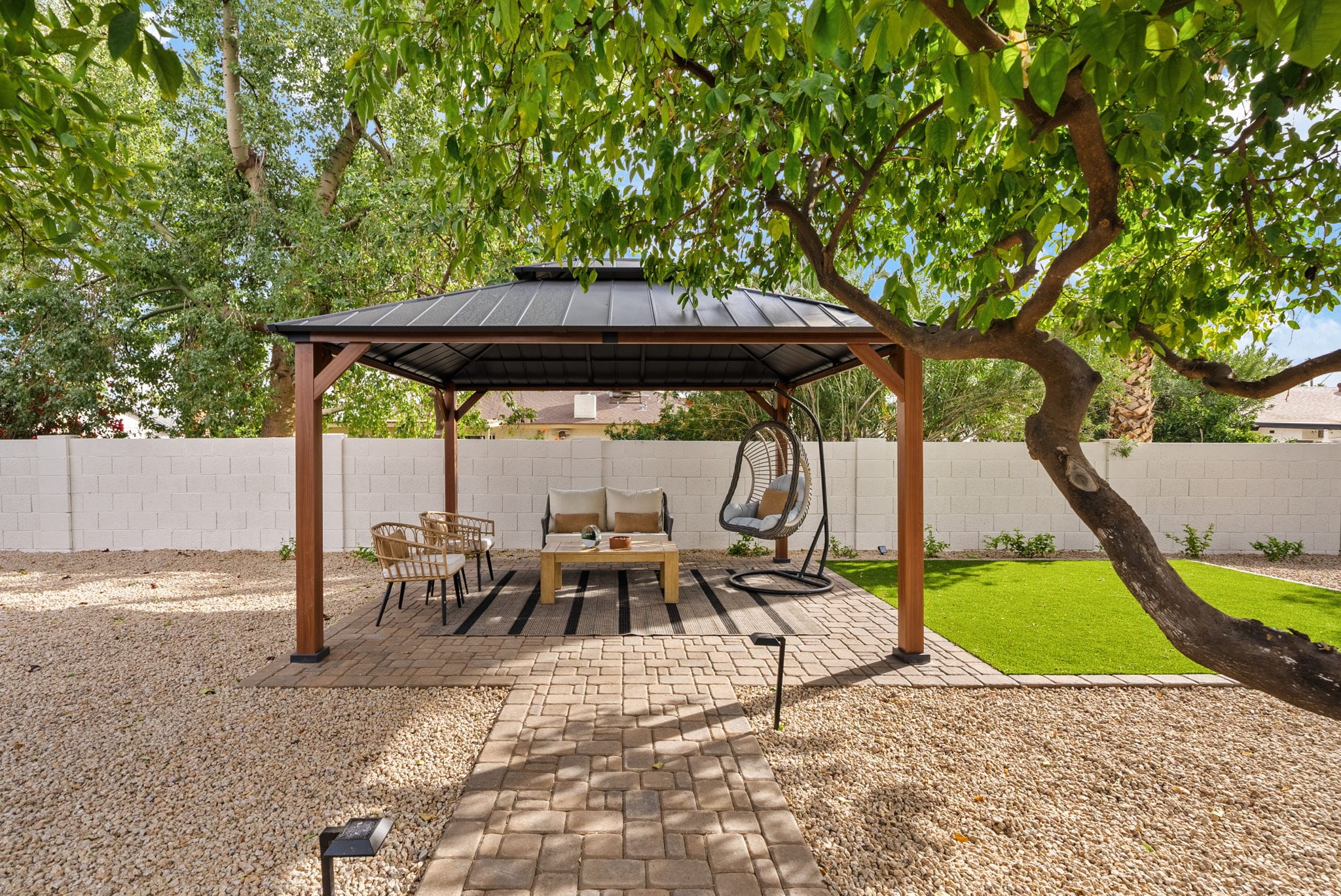 A shaded backyard features a dark metal gazebo with a sofa, chairs, and a hanging egg chair on a paved patio, surrounded by gravel, artificial grass, and a white block wall.