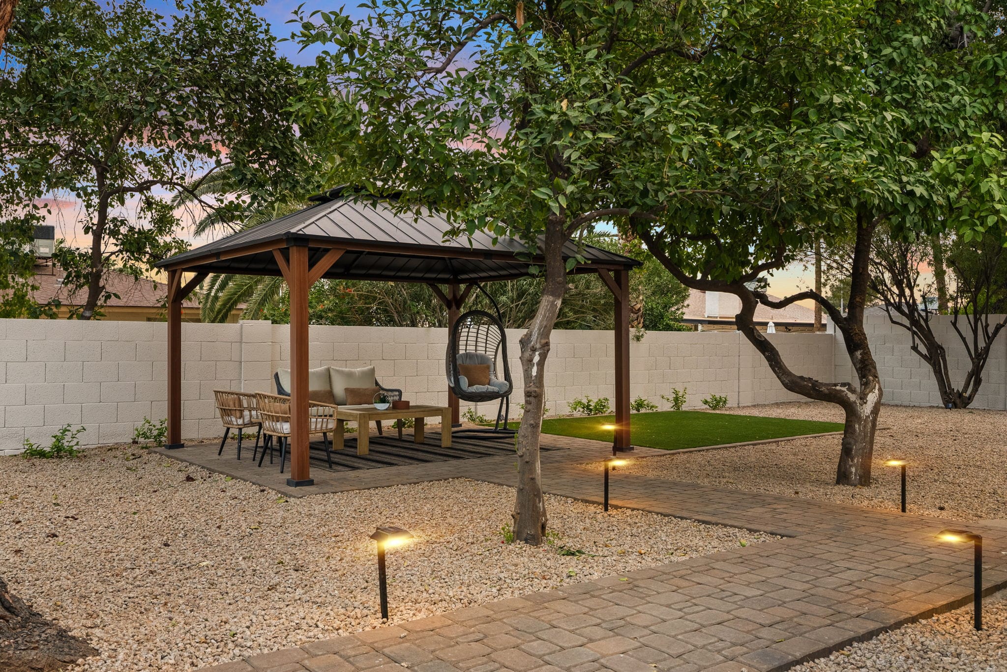 A landscaped backyard at twilight featuring a gazebo with outdoor seating, a paved pathway illuminated by lights, gravel, and several trees against a white block wall.