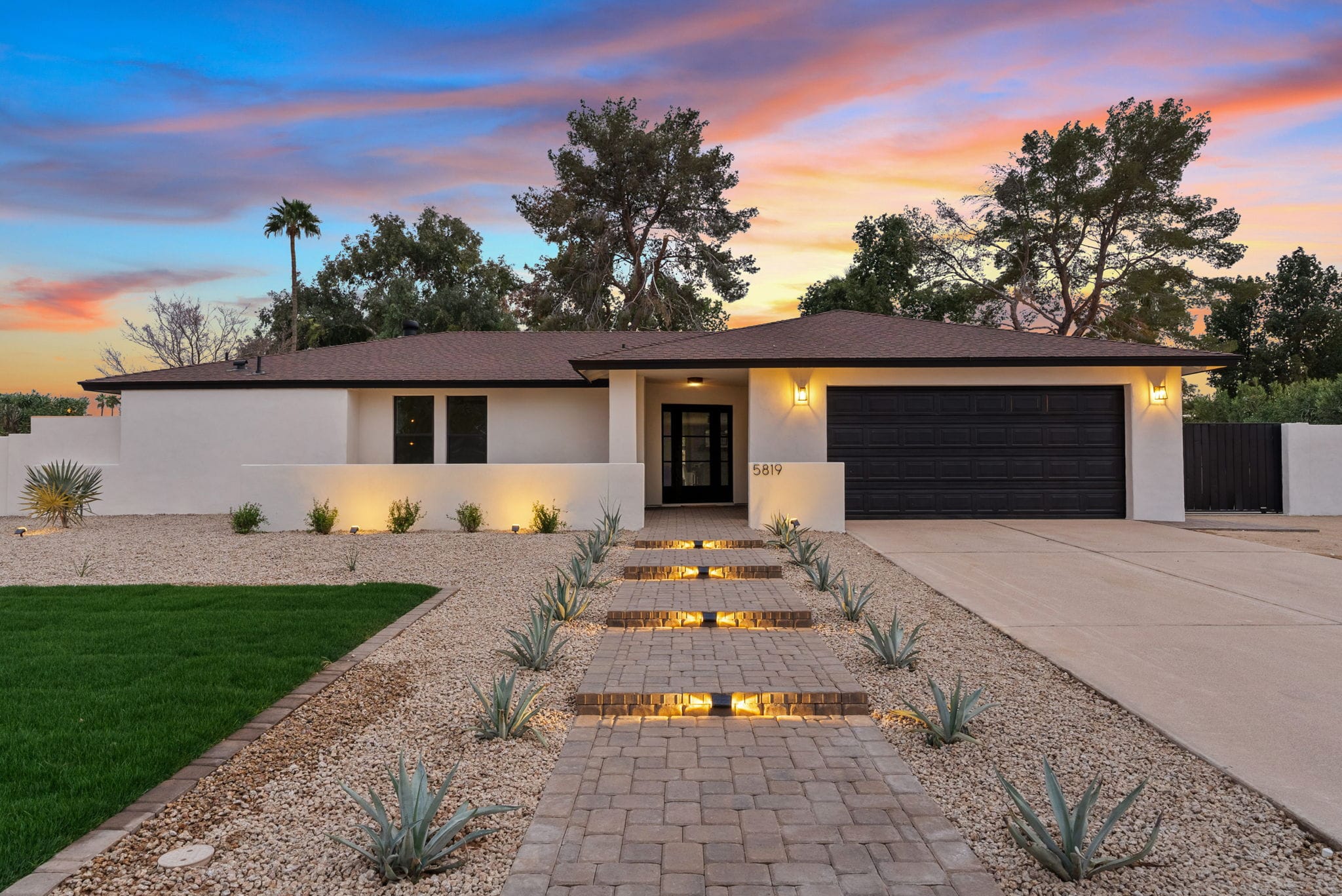 A modern white house with a black front door and garage at dusk, featuring a lighted paver walkway, agave plants, gravel, green lawn, and a vibrant sunset sky.