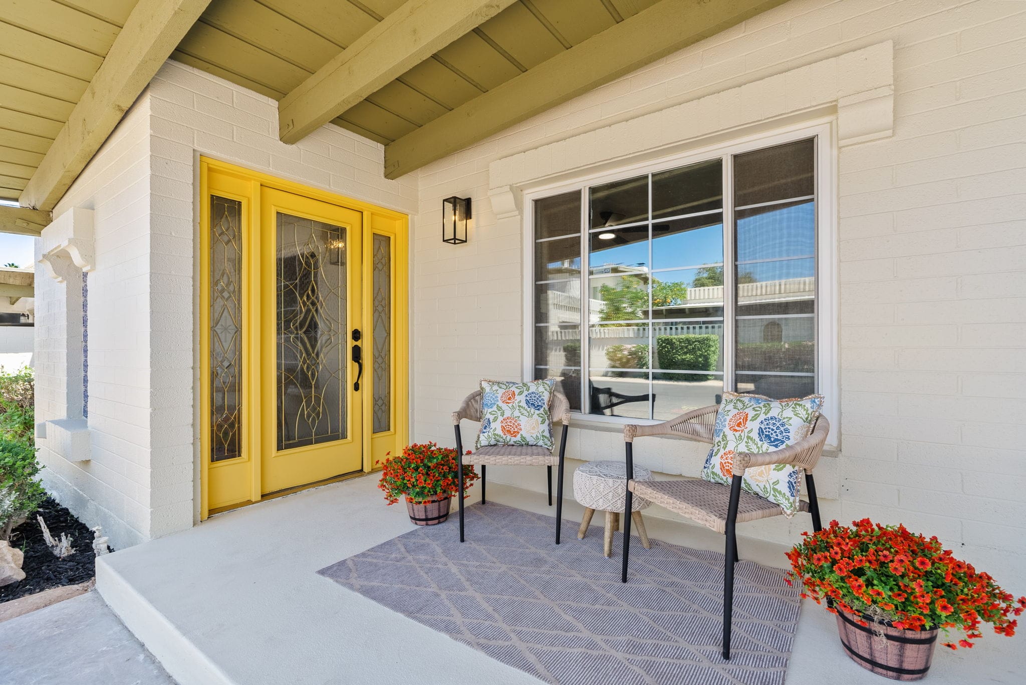A bright yellow front door and white brick porch with two wicker chairs, floral pillows, red potted flowers, and a patterned rug.