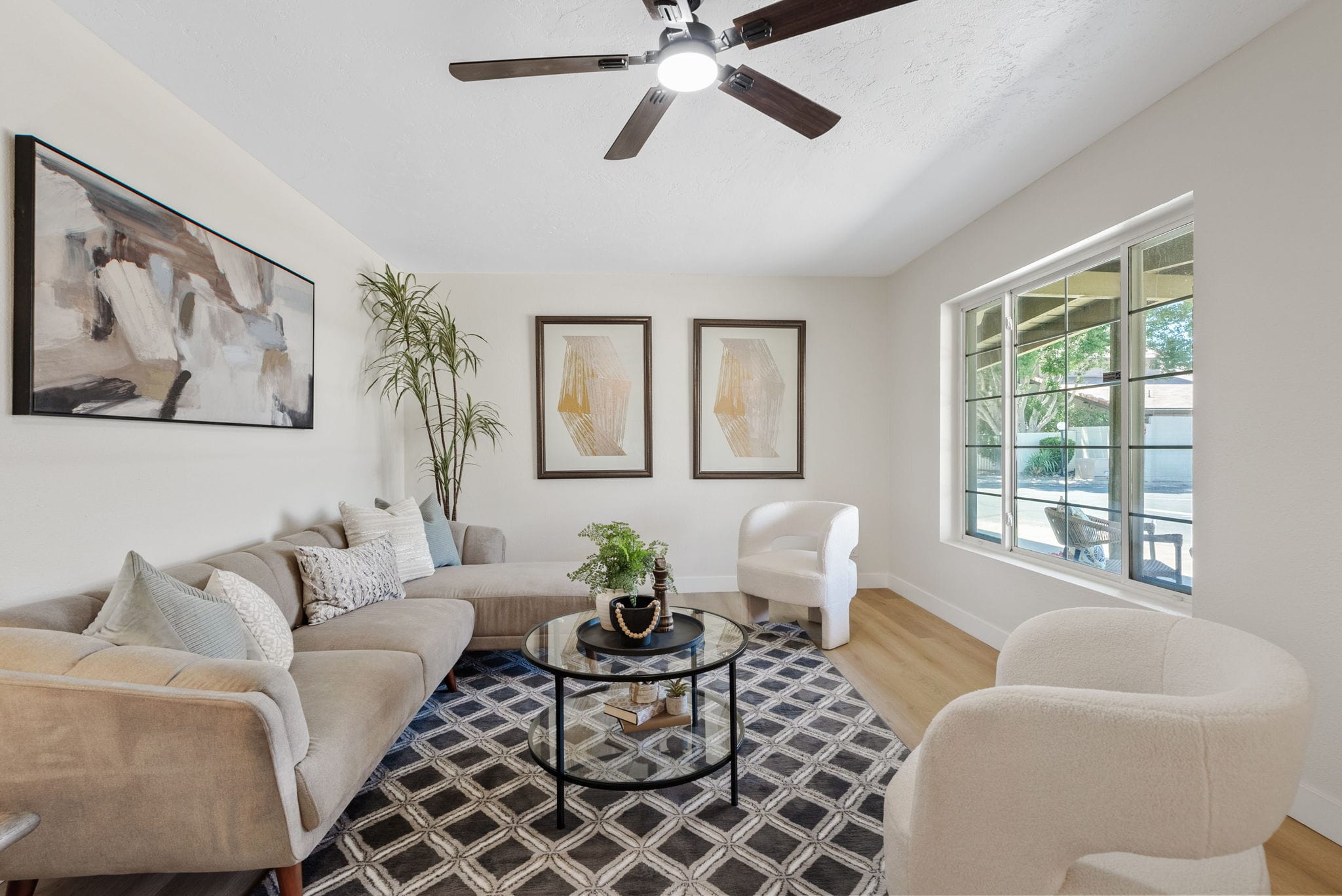 A bright, modern living room featuring a light sectional sofa, two white textured chairs, a glass coffee table, geometric rug, abstract art, and a large window.