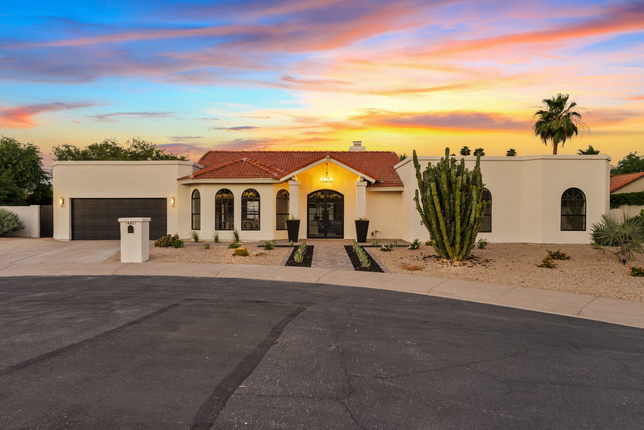 Mediterranean-style house with red tile roof, light stucco, arched windows, and an illuminated entrance, set against a vibrant sunset sky with desert landscaping.