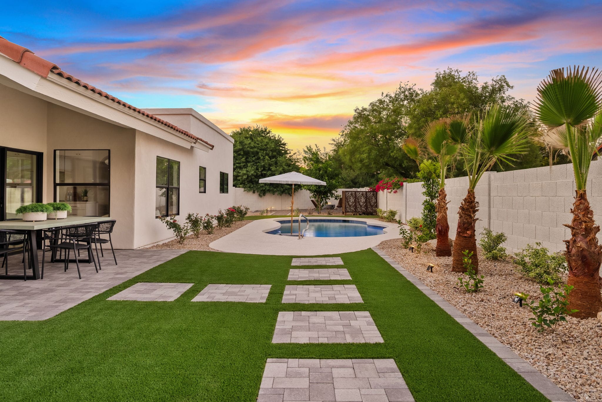 A spacious backyard at sunset featuring a white house, outdoor dining patio, artificial turf with stepping stones, a swimming pool, and desert landscaping with palm trees.