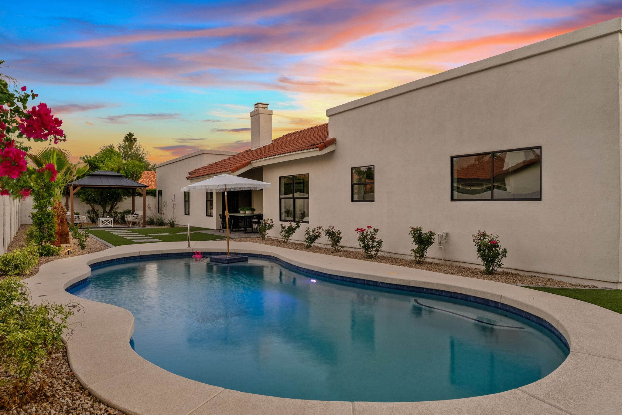 A modern backyard features a curved swimming pool, a stucco house with a tile roof, an outdoor dining area, and a gazebo, all illuminated by a vibrant sunset sky.