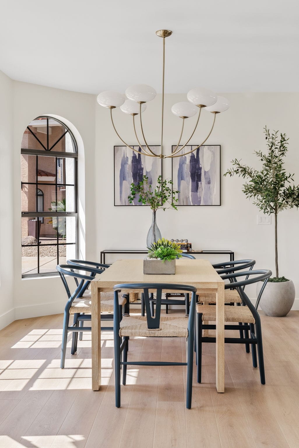 Modern dining room with a light wood table, dark blue wishbone chairs, a brass chandelier, abstract art, and potted plants, illuminated by an arched window.
