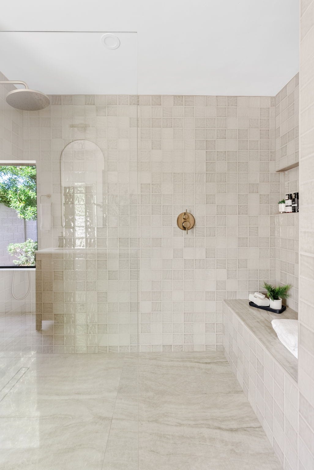 A modern, spacious shower with light-colored square tiles, a large rain shower head, a golden control knob, and a built-in bench with towels and plants, next to a window.