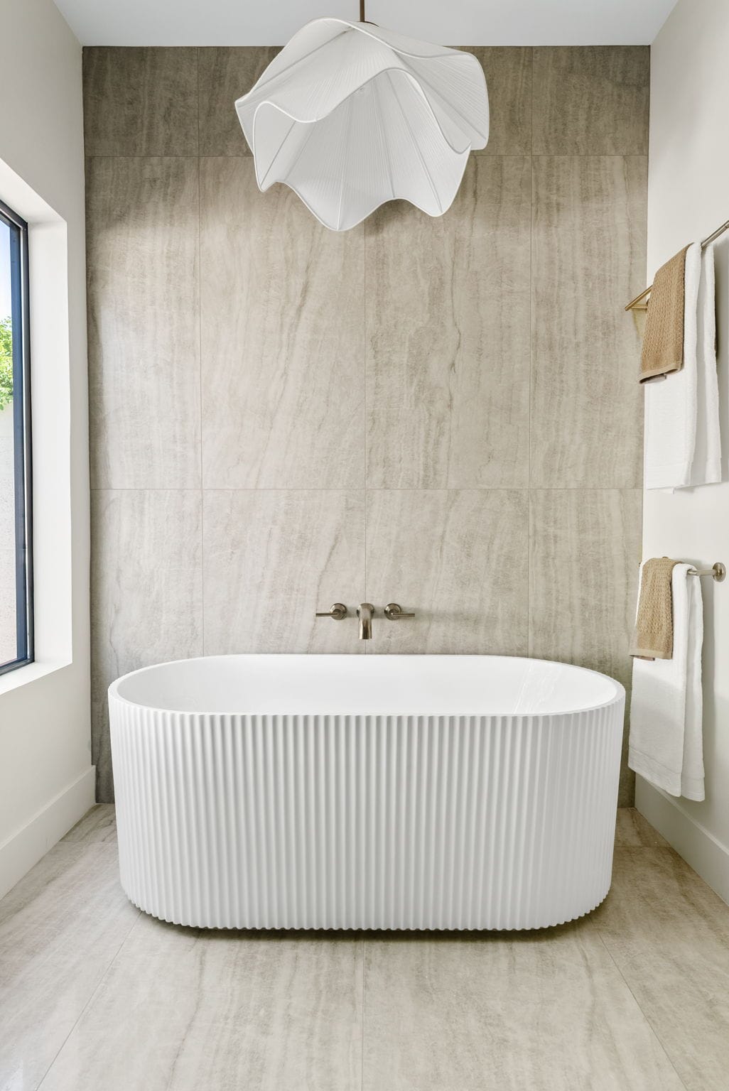 Modern bathroom with a white fluted freestanding tub against light marble-effect tiled walls. A wavy pleated pendant light hangs above, with a wall-mounted faucet and towels on racks.