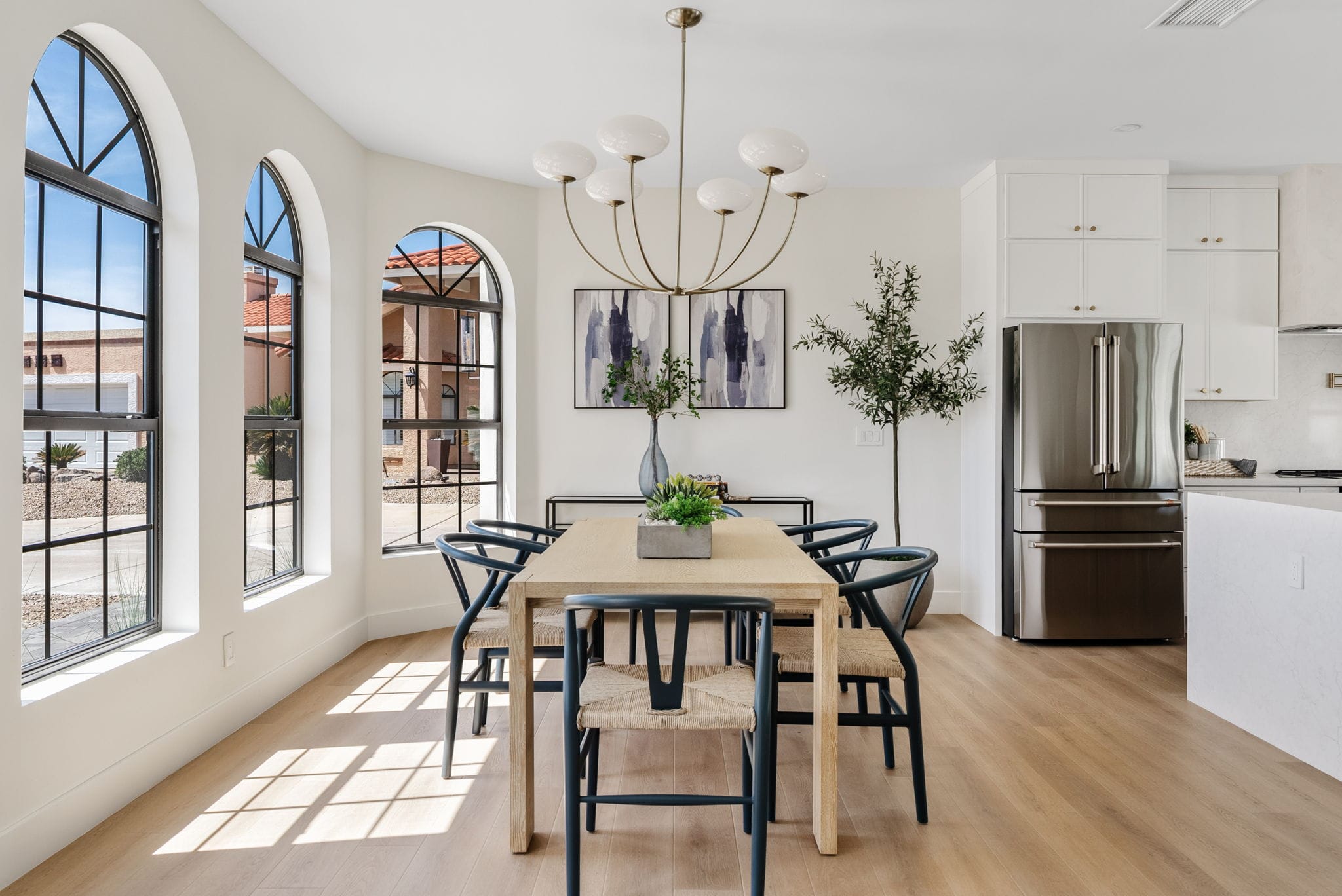 A bright, modern open-concept dining room and kitchen. The dining area features a light wood table, dark chairs, and arched windows, while the kitchen has white cabinets and a stainless steel refrigerator.