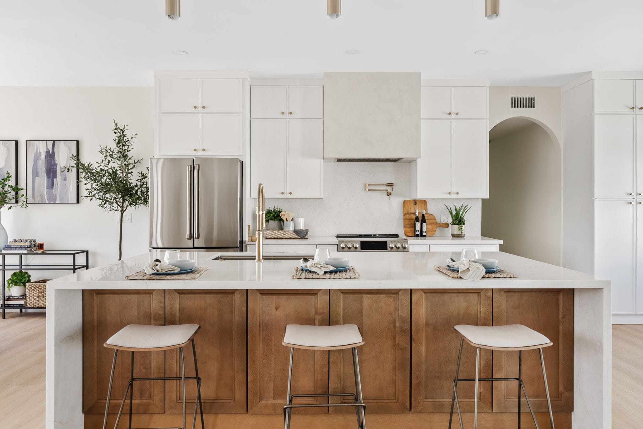 A bright, modern kitchen with white upper cabinets, a large island with natural wood base cabinets and white countertop, stainless steel appliances, gold accents, and three bar stools.