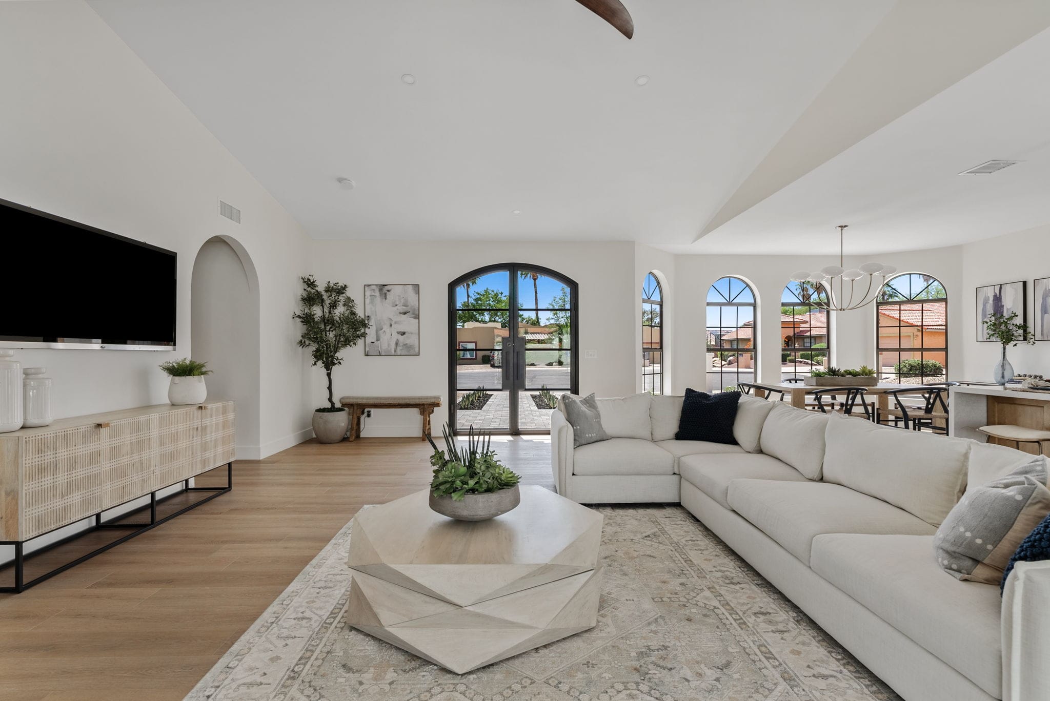 A bright, open-concept living room with a large white sectional sofa, unique geometric coffee table, TV console, and arched windows looking out to a sunny neighborhood.