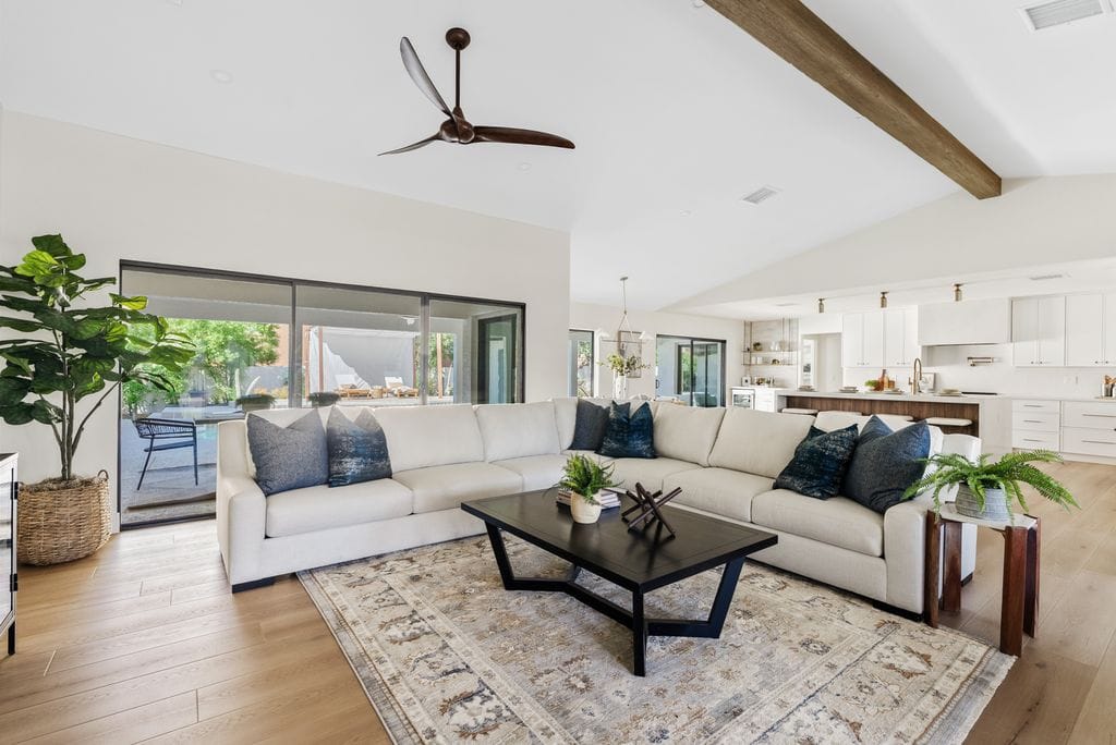 Bright, modern open-concept living area featuring a large white sectional, dark coffee table, and patterned rug, seamlessly connecting to a white kitchen and outdoor patio.